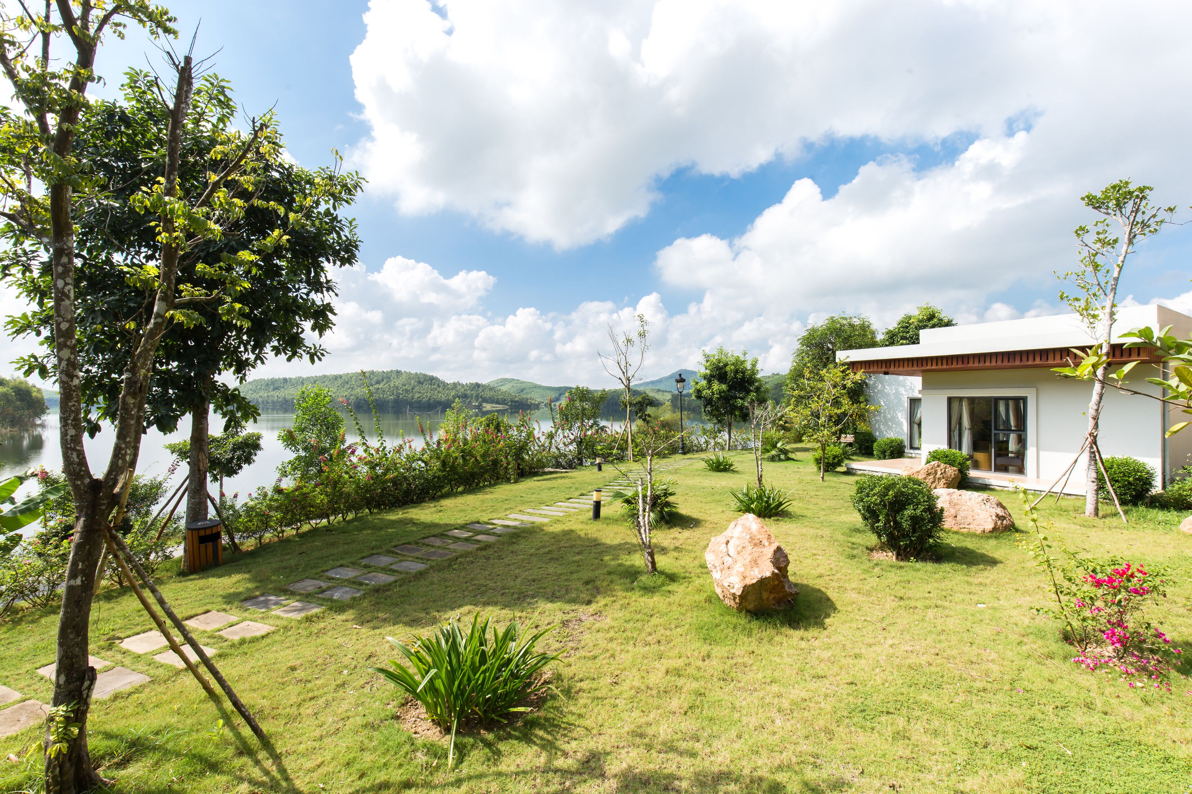 A modern house with a stone pathway leading to a door, surrounded by lush greenery and a lake view.