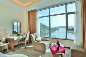 A hotel room with a large window offering a scenic view of a lake and mountains, a desk with a computer, a chair, and a small table with a wine bottle and glass. - Muong Thanh Luxury Dien Lam Hotel (Hung Chau)