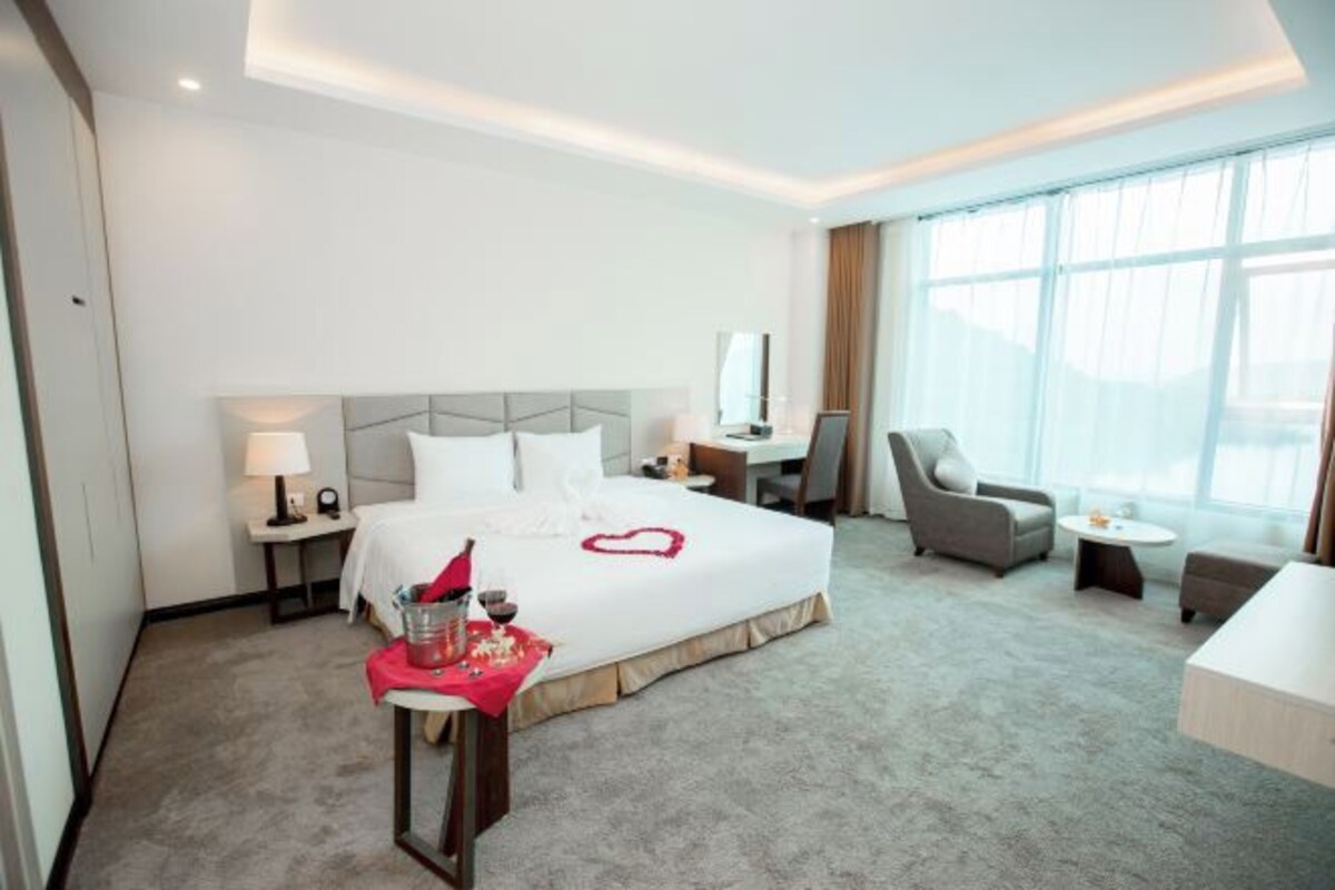 A hotel room with a large bed, a desk, a chair, and a small table with a drink and a heart-shaped decoration.