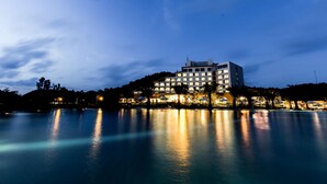 A modern hotel with multiple floors, located near a body of water, with lights reflecting on the water surface. - Muong Thanh Luxury Dien Lam Hotel (Hung Chau)