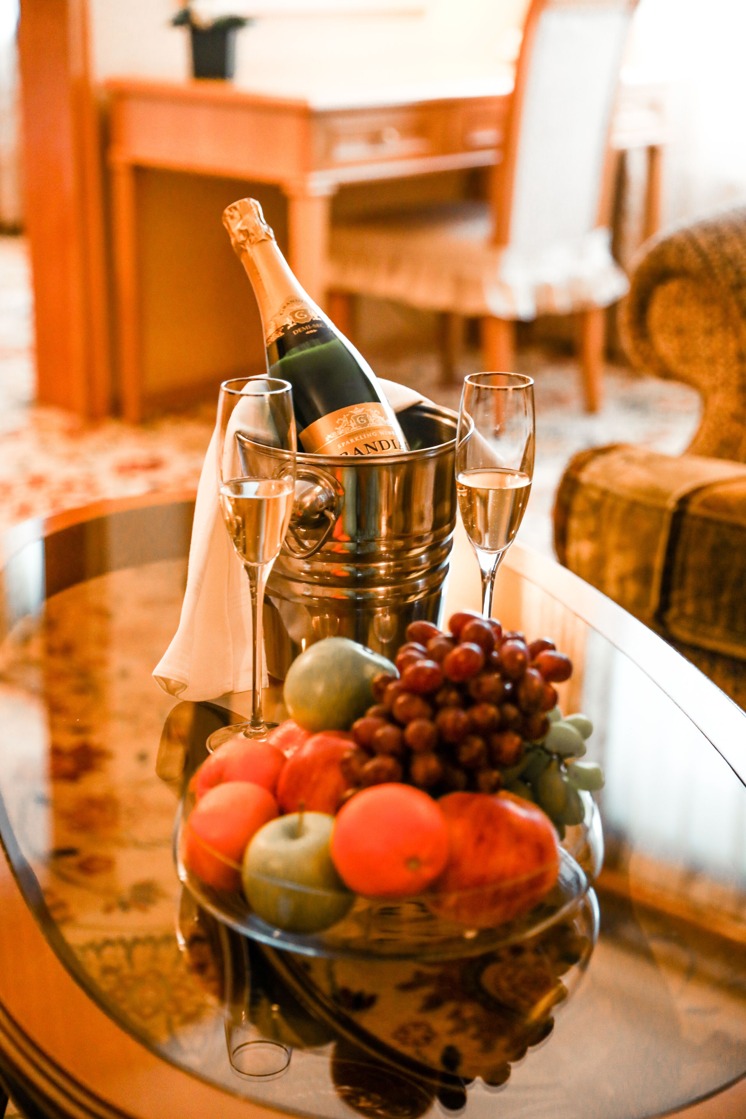 A glass table with a bottle of champagne, two champagne glasses, and a bowl of fruit.