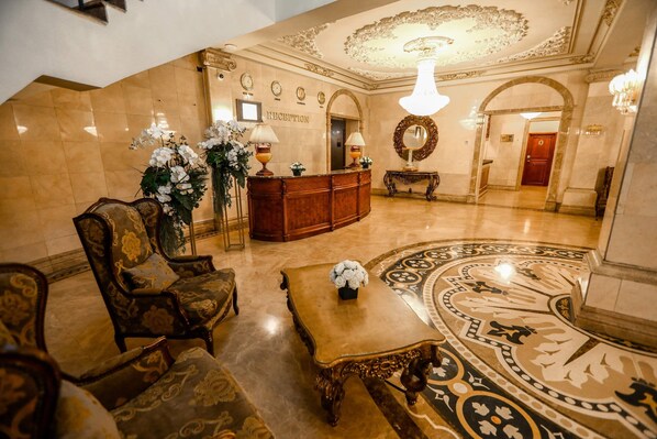 A lobby with a reception desk, ornate chairs, a coffee table, and a decorative floor pattern.