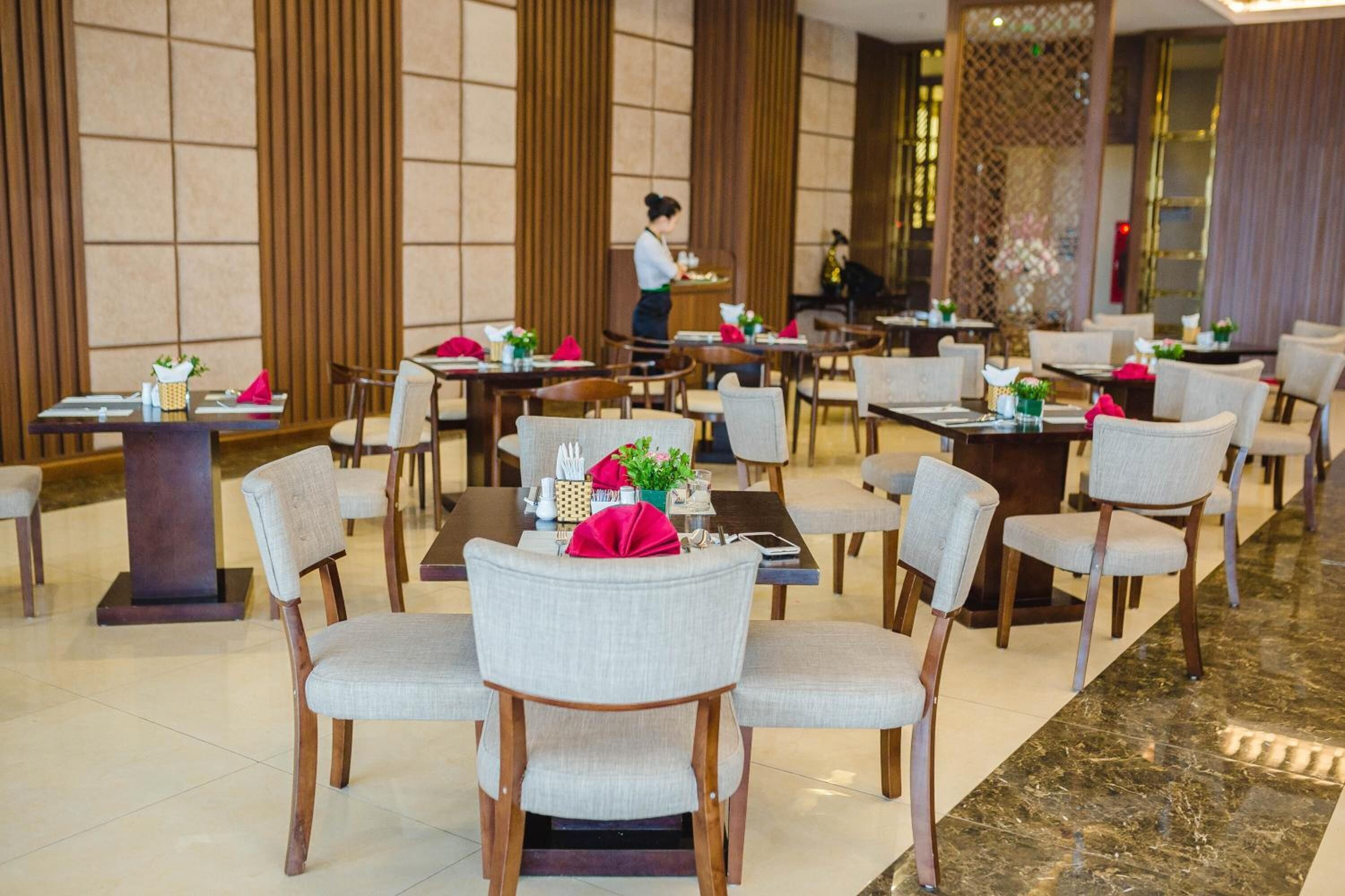A restaurant with multiple tables and chairs, a waiter attending to a table, and a decorative wall in the background.