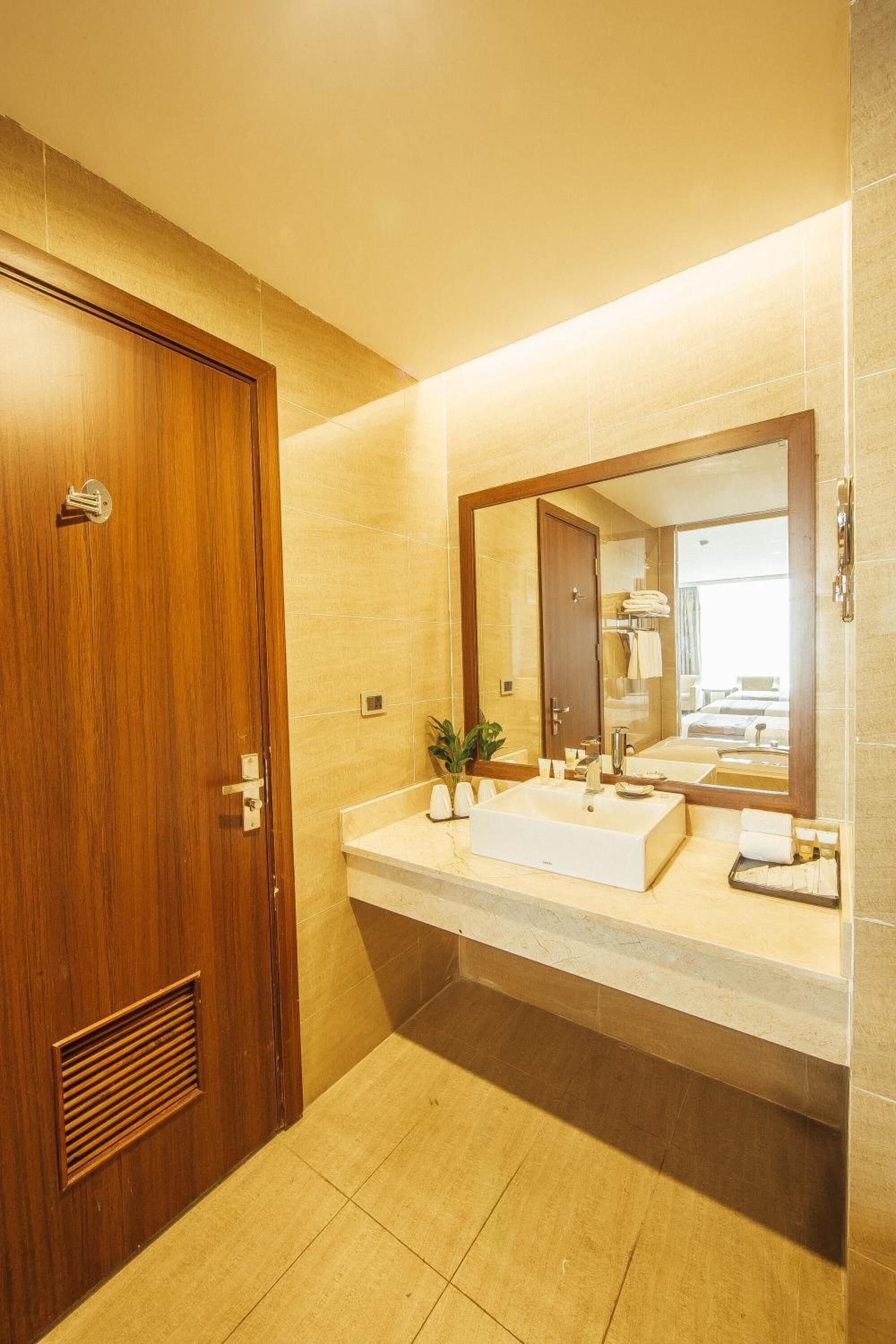 Un baño con puerta de madera, un espejo grande, un lavabo blanco y piso de baldosas.
