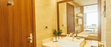 Un baño con puerta de madera, un espejo grande, un lavabo blanco y piso de baldosas.
