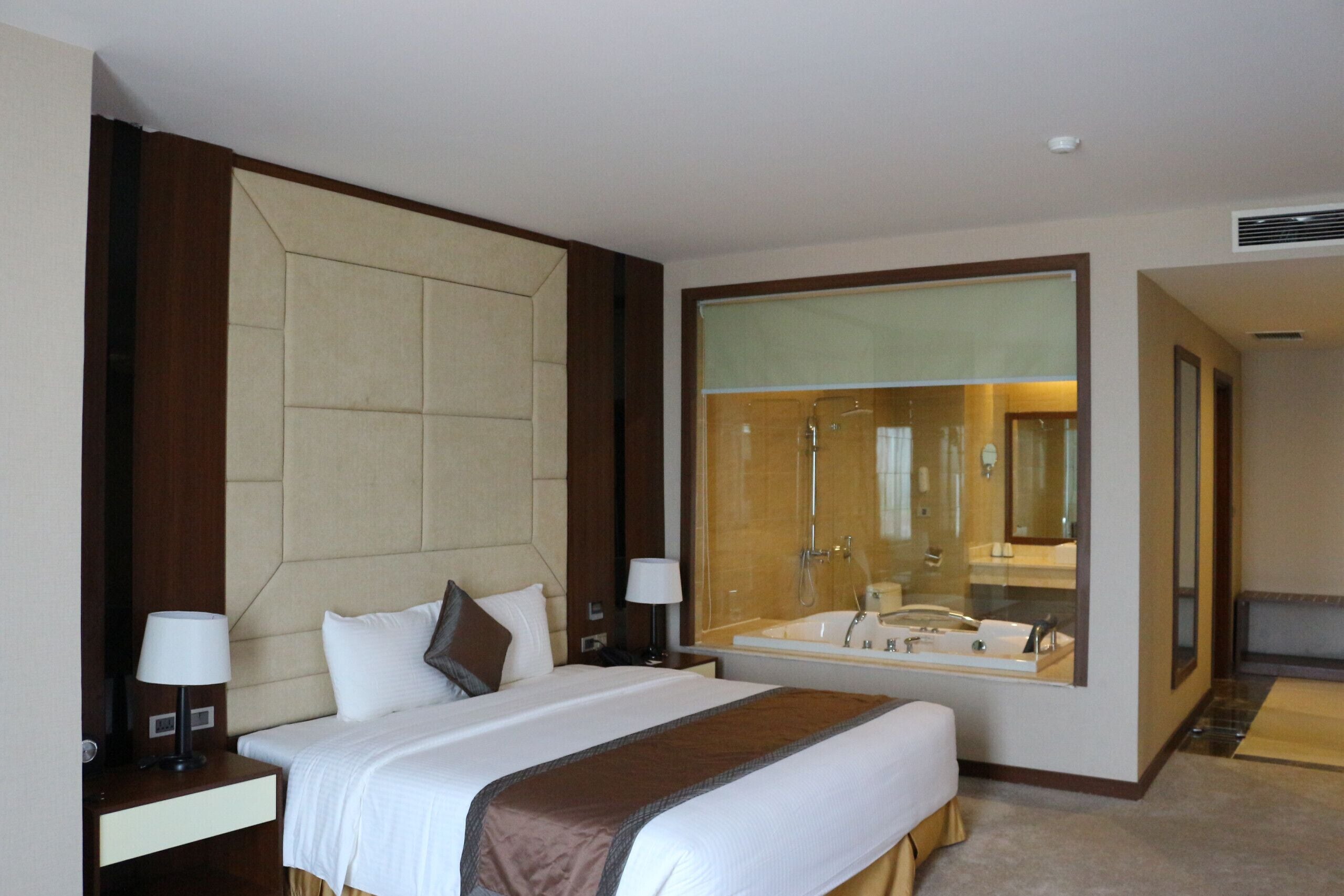 A hotel room with a large bed, a nightstand, a lamp, a bathroom with a bathtub, and a large mirror.