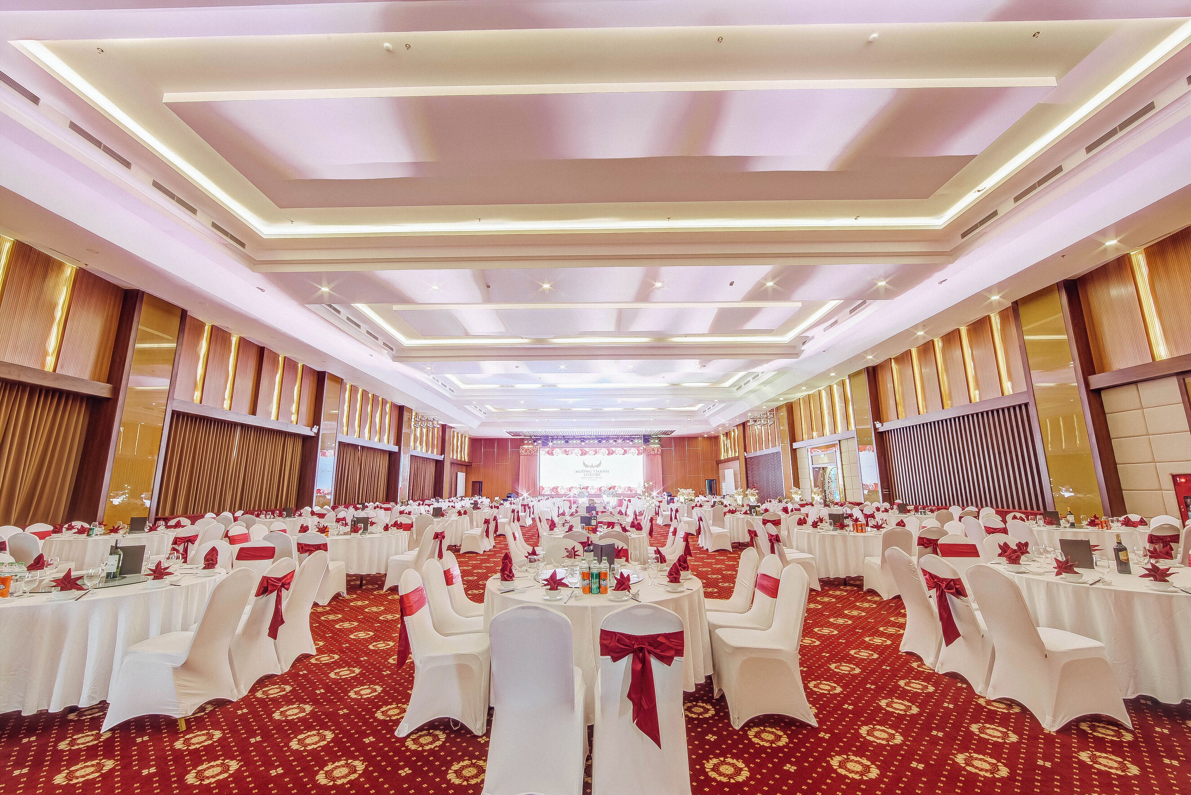A banquet hall with round tables, white chairs with red sashes, and a red carpeted floor.