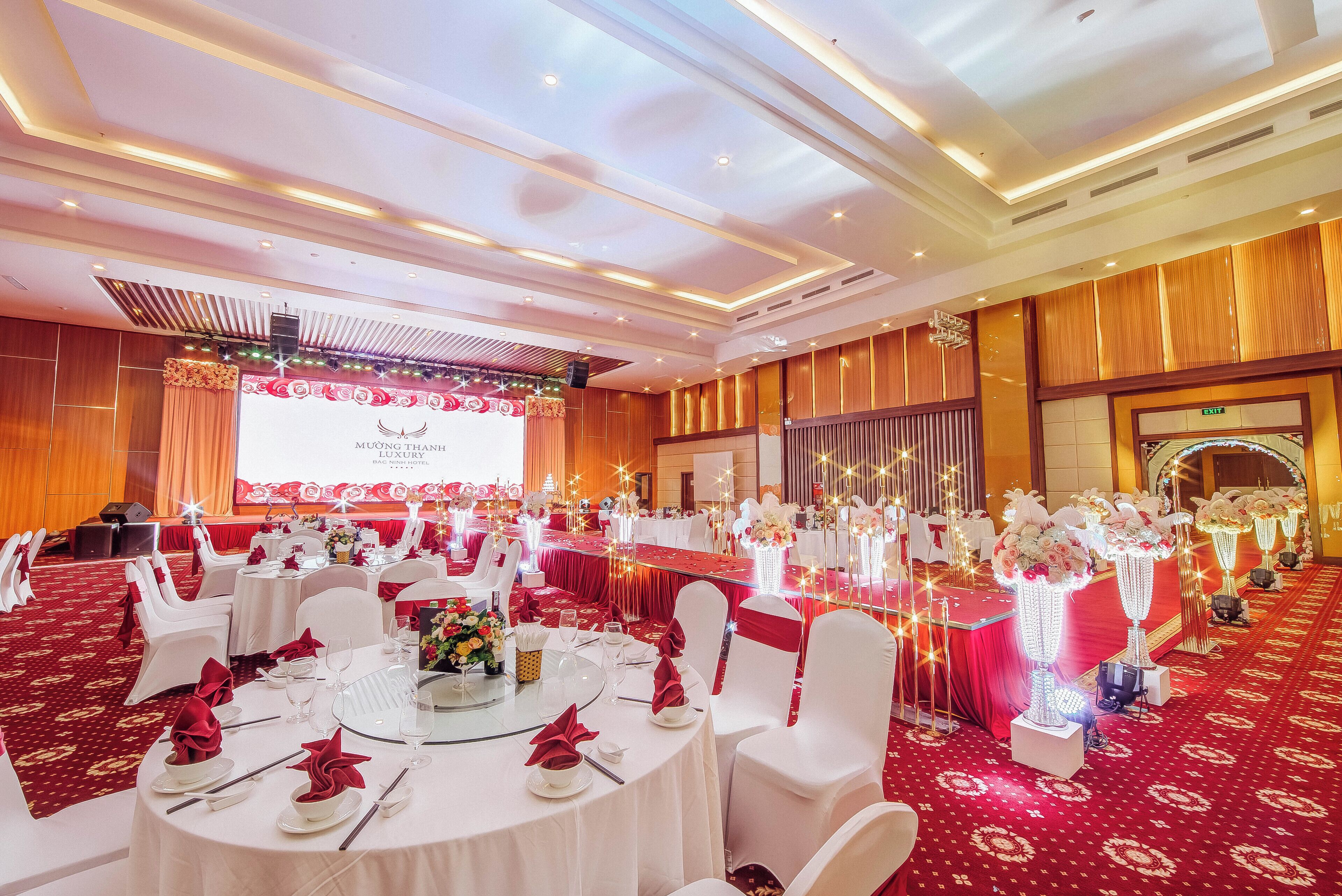 A banquet hall with round tables set for dining, white chairs, and a stage with a red backdrop.