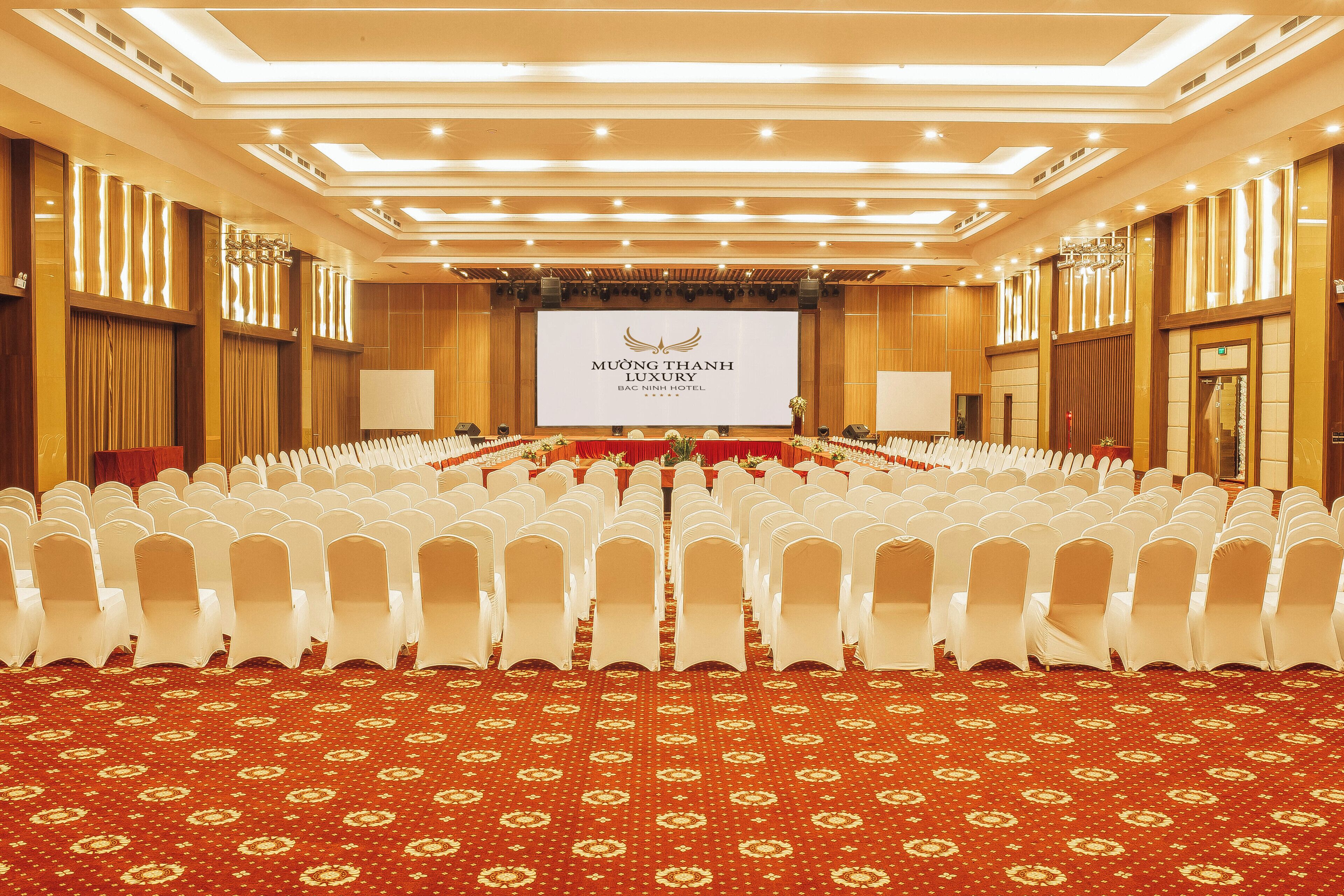 Uma sala de conferências com fileiras de cadeiras brancas, um tapete vermelho e uma tela grande exibindo o nome do hotel.