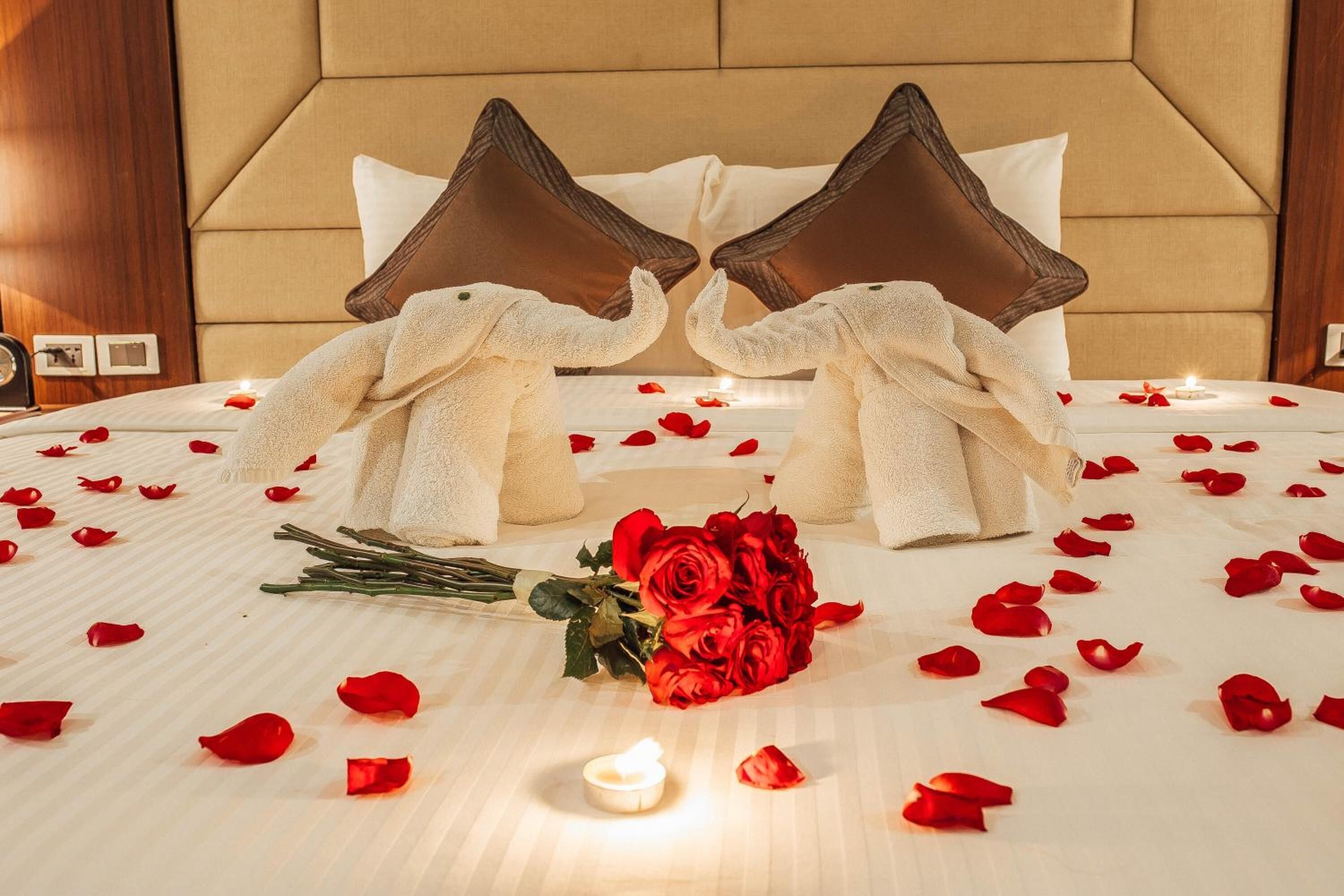 Una cama con pétalos de rosa y una vela, dos toallas con forma de cabeza de elefante y un ramo de rosas rojas.
