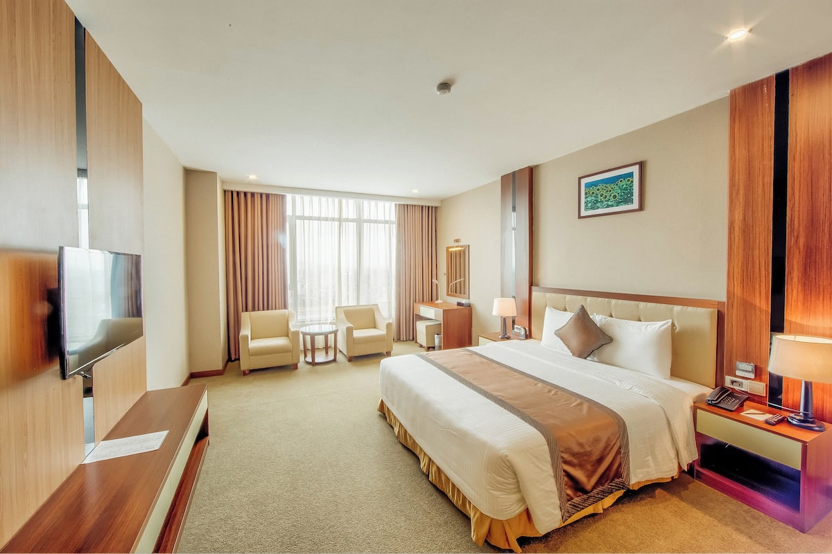 A hotel room with a large bed, a desk, a TV, a chair, a small table, and a window with curtains.