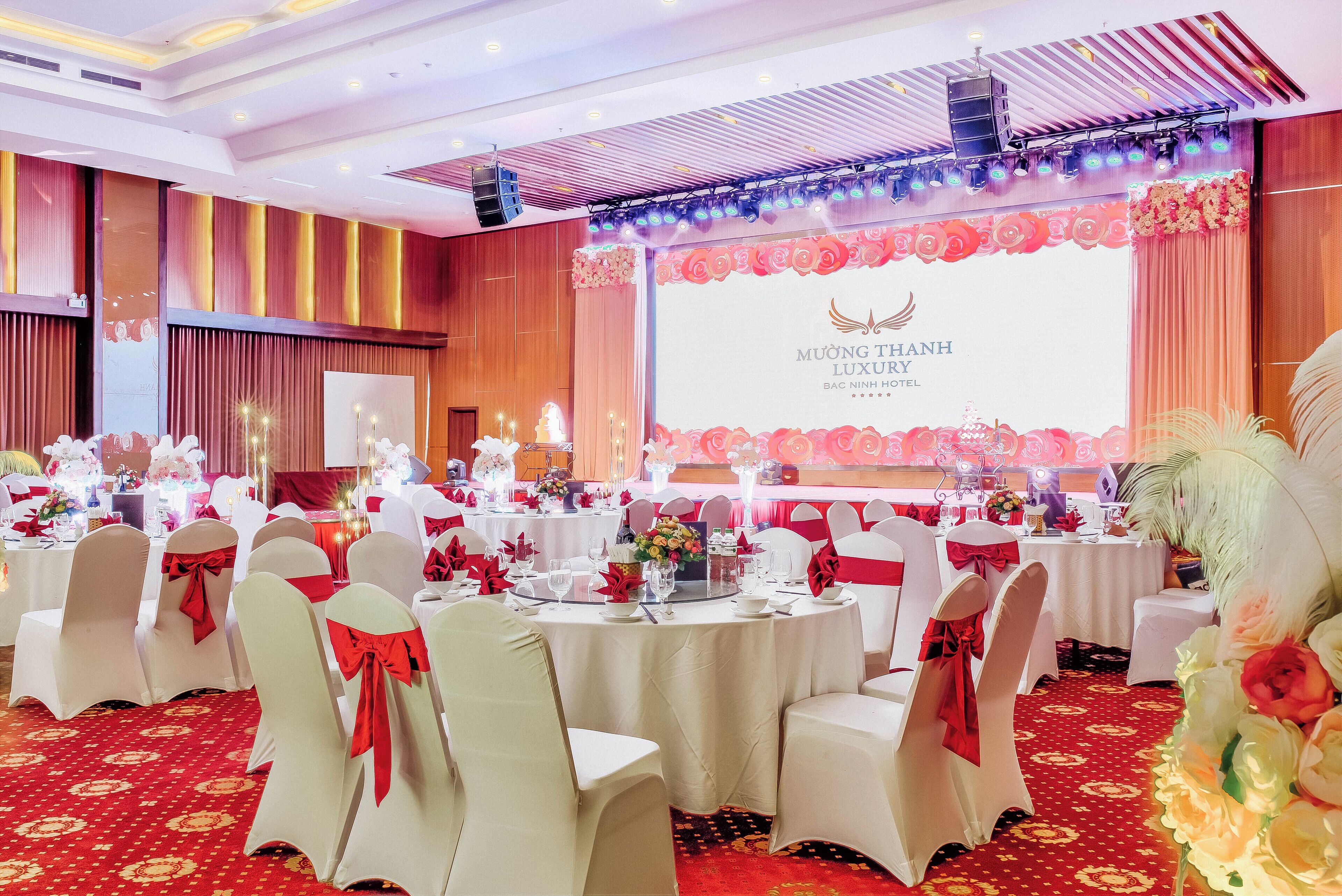 A banquet hall with round tables, white chairs with red sashes, and a stage with a backdrop for MUONG THANH LUXURY BANC NHINH HOTEL.