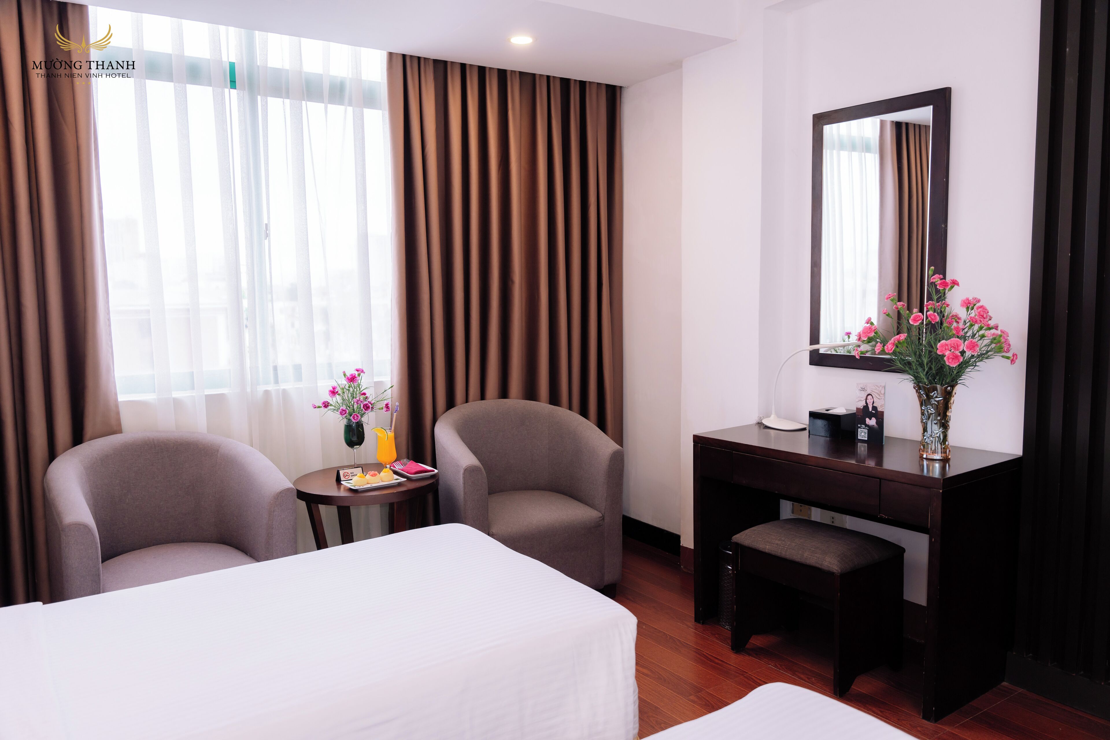 A hotel room with two beds, a desk, a chair, a small table with a vase of flowers, and a large mirror.