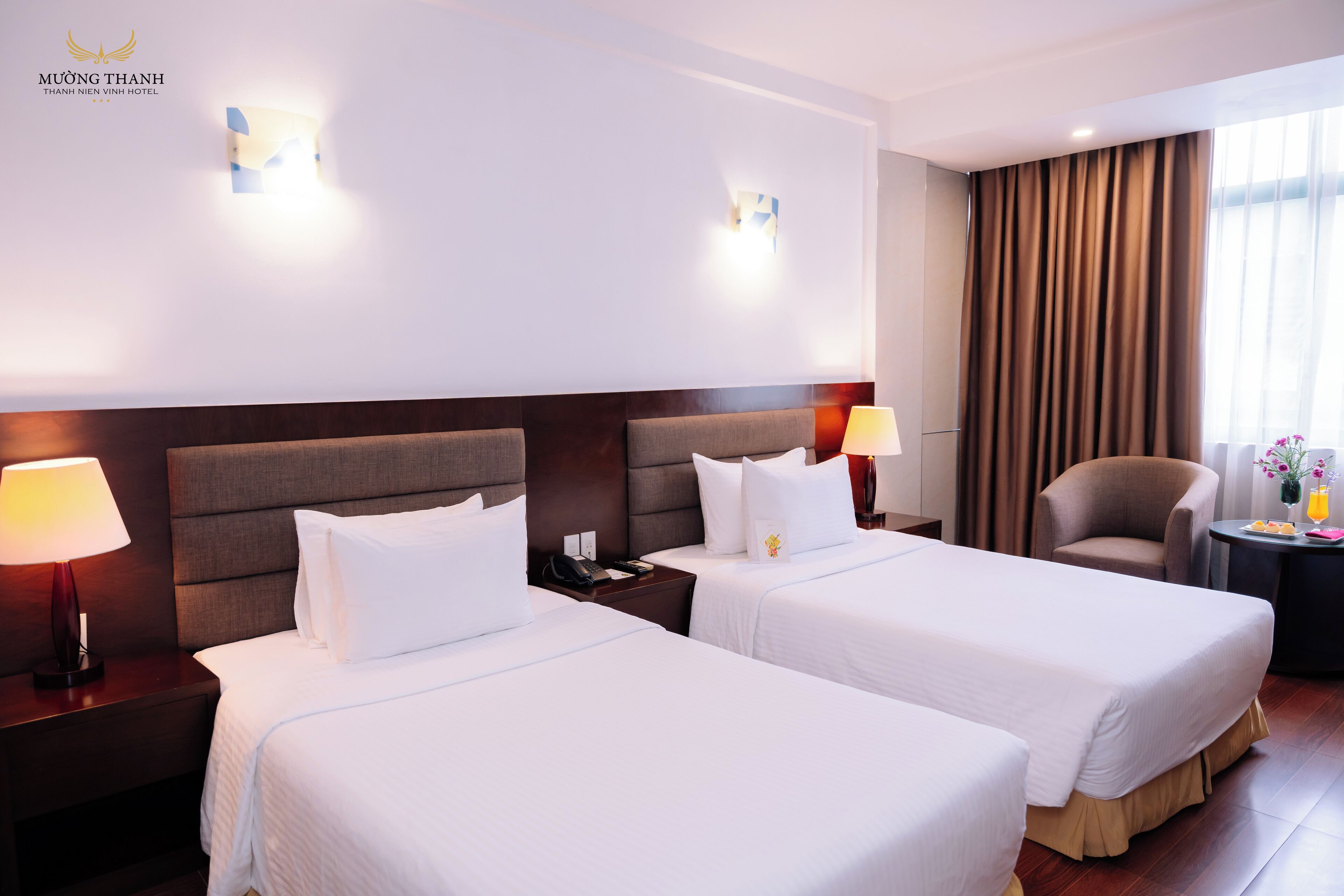 Habitación de hotel con dos camas, una mesita de noche con lámpara, una silla y una pequeña mesa con un florero y fruta.