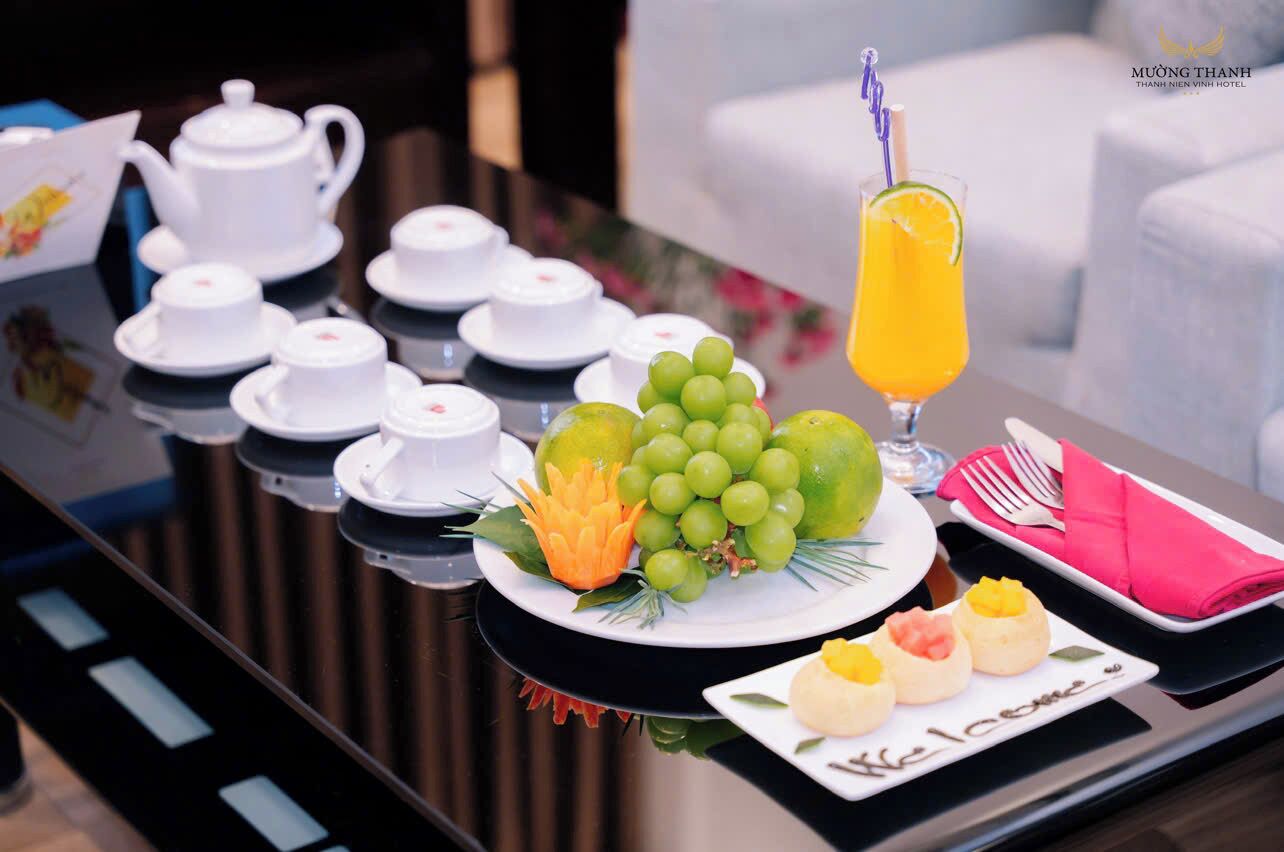 A breakfast setup with white teapots, cups, a plate of green grapes, a carved orange, and a glass of orange juice.