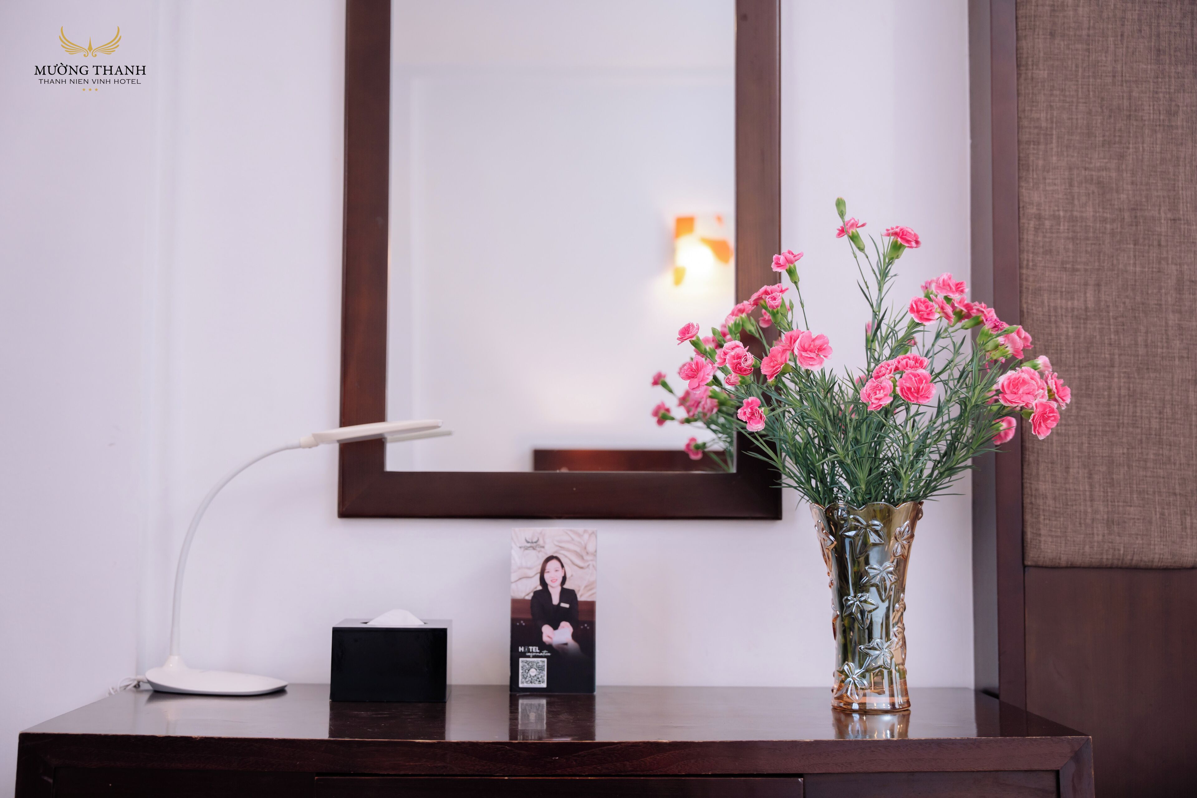 A hotel reception desk with a framed photograph, a vase of pink flowers, and a desk lamp.