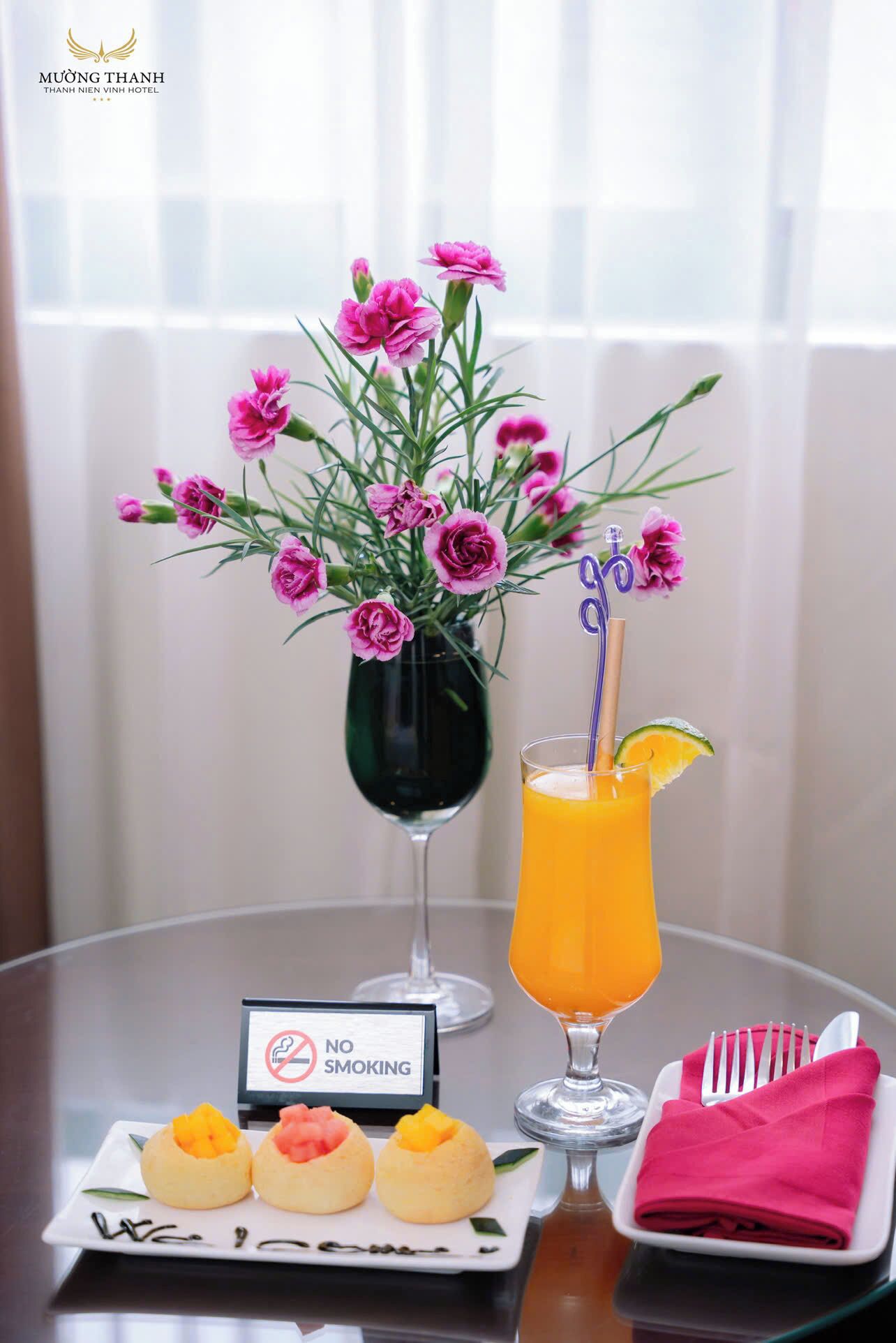 A table with a vase of pink flowers, a glass of orange juice with a straw, and a plate of fruit. A 'No Smoking' sign is visible.