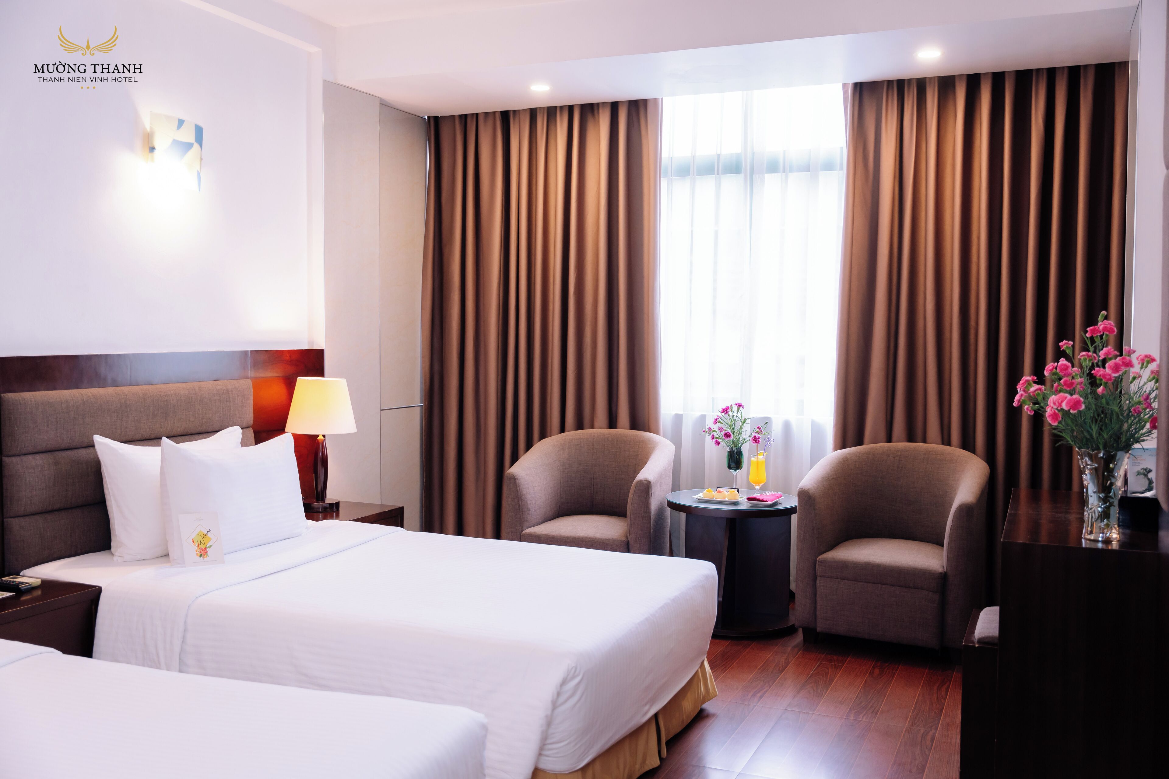 Habitación de hotel con dos camas, una zona de estar con una silla, una mesita con un jarrón de flores y una mesita de noche con una lámpara.