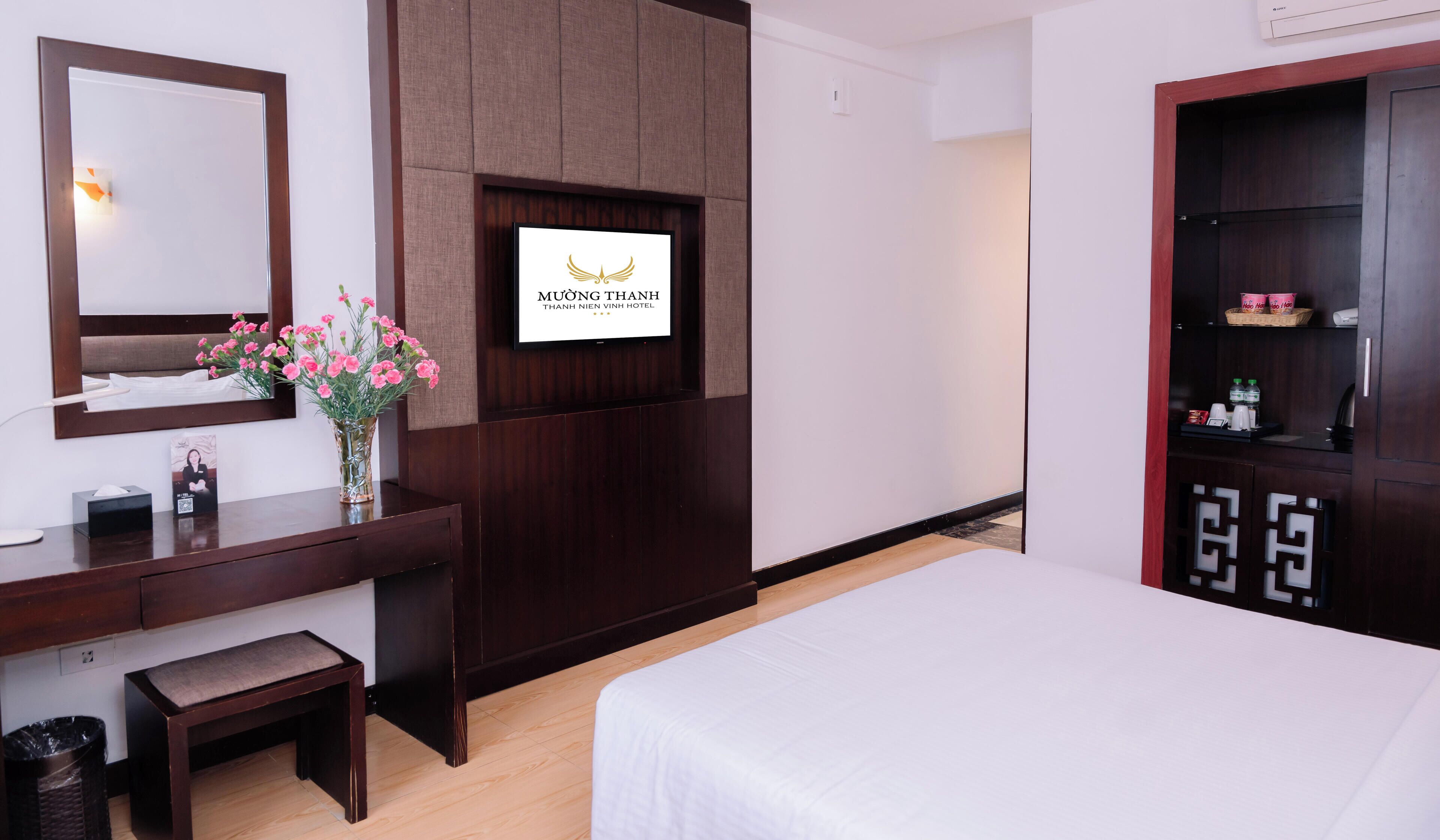 Habitación de hotel con una cama, un escritorio con espejo, un florero y un televisor de pared que muestra el nombre del hotel.