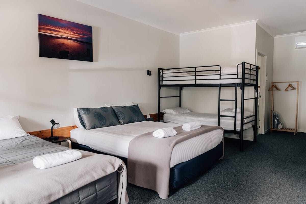 A hotel room with two single beds, a bunk bed, a desk with a lamp, a coat rack, and a painting of a sunset.