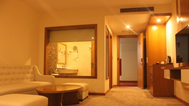 Une chambre d’hôtel comprenant un canapé, une table basse ronde, une petite table avec une lampe, une salle de bain avec un lavabo et un miroir, et un placard.
