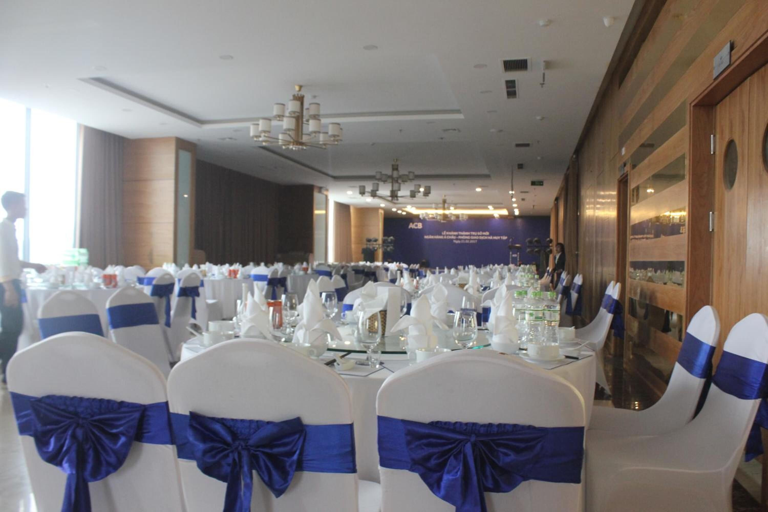 A banquet hall with rows of white chairs with blue sashes, tables set for a formal event, and a large banner with text in the background.