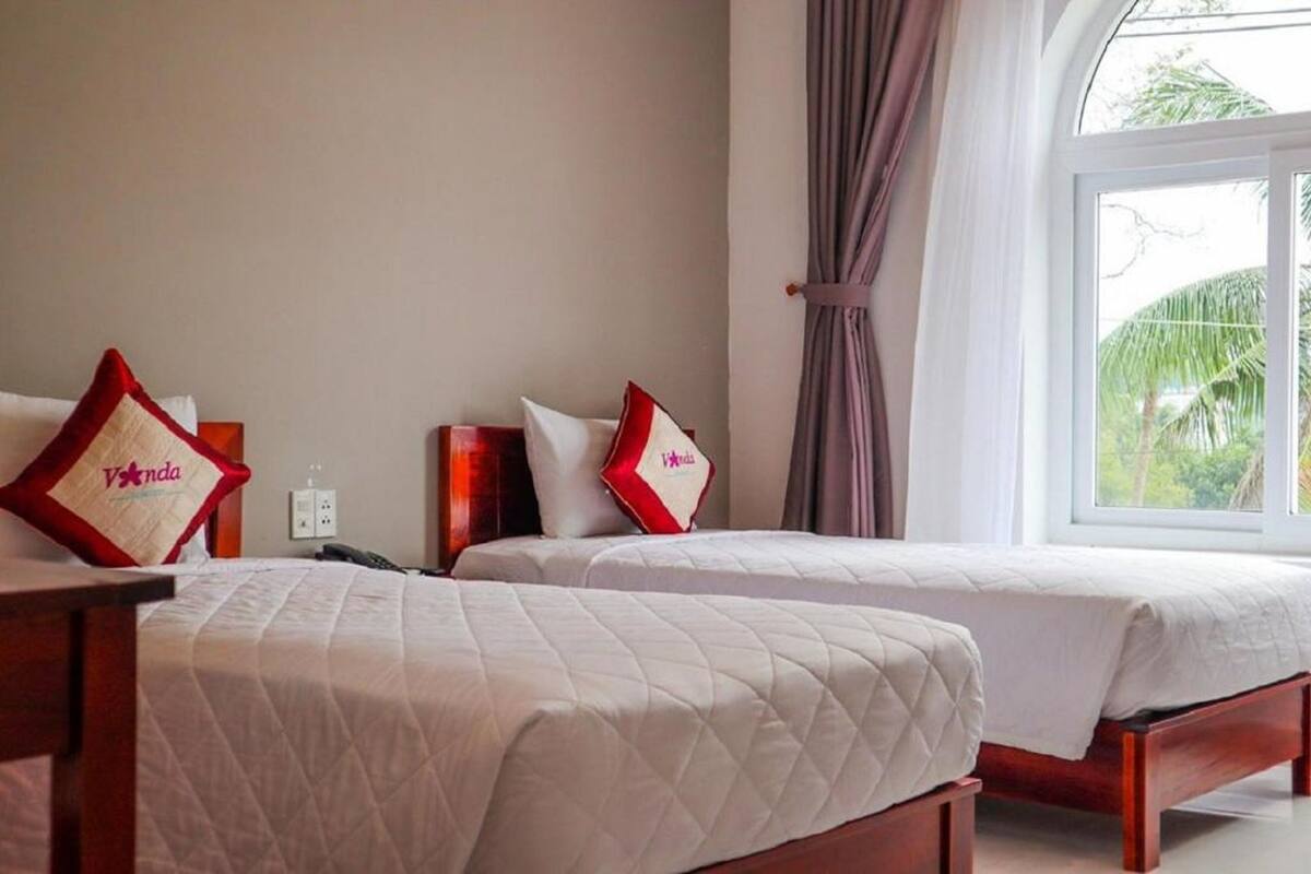 Two beds with white bedding and red pillows with 'Vanda' branding, a window with curtains, and a view of greenery outside.