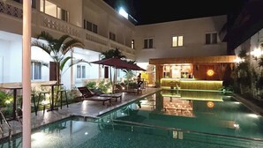Ein Poolbereich mit Liegestühlen und Sonnenschirmen, ein Barbereich mit einer Neonreklame und ein Gebäude mit zahlreichen Fenstern.