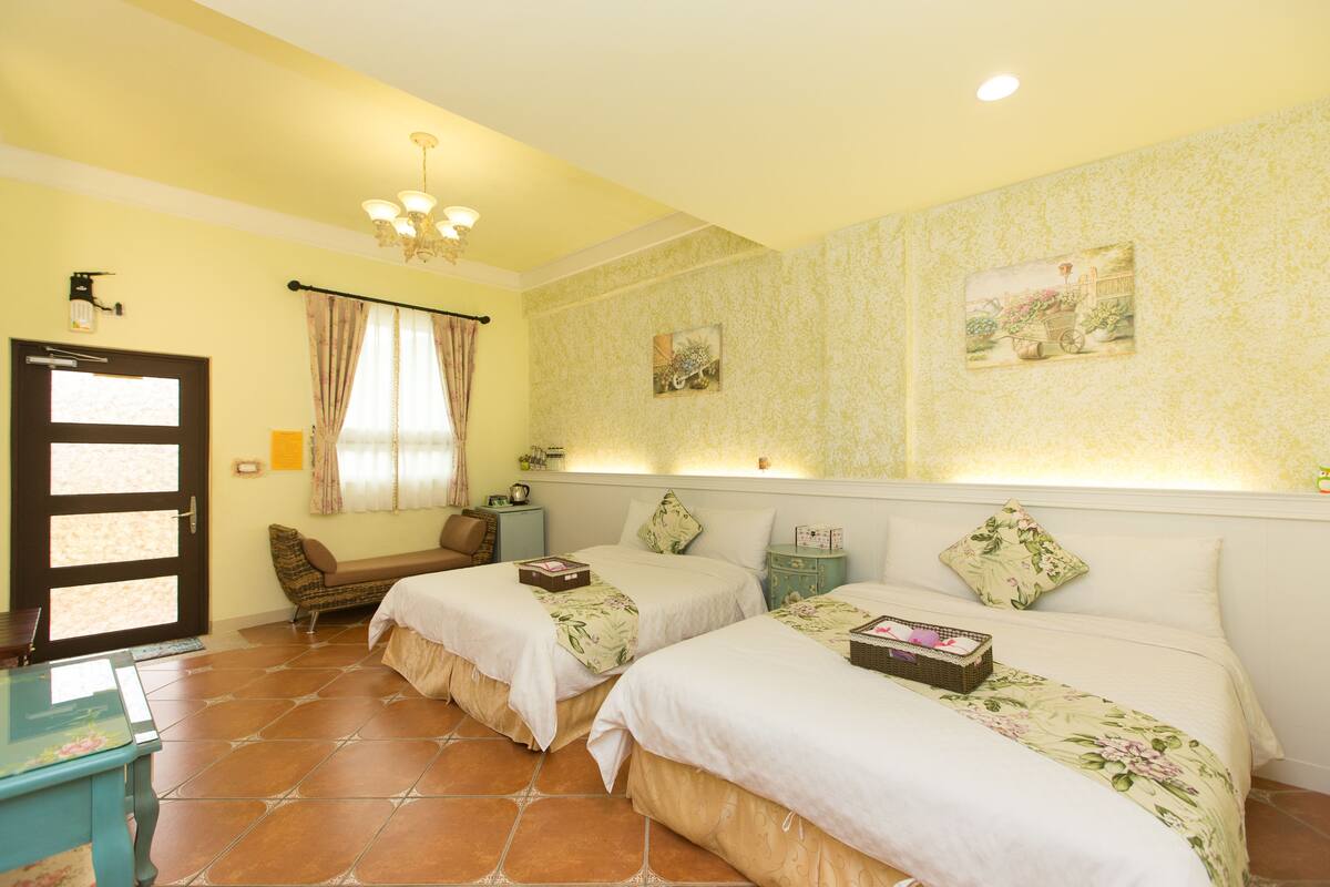 A hotel room with two beds, a bench, a small table, and a door.