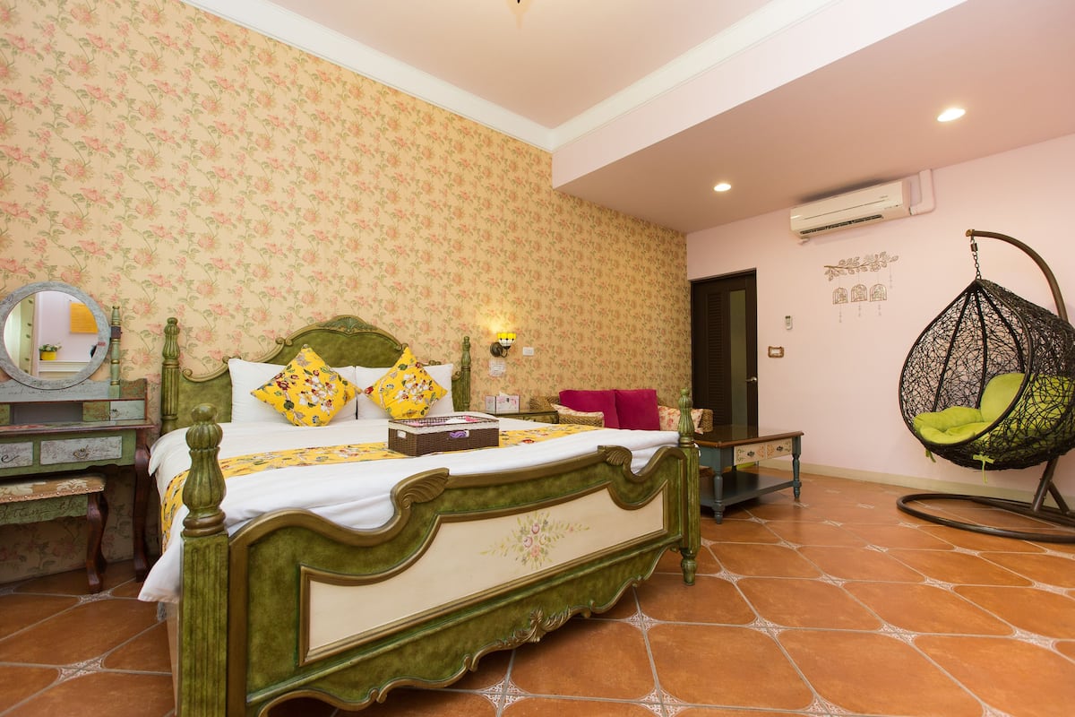 A bedroom with a green bed, floral wallpaper, a hanging chair, and a bedside table with a mirror.