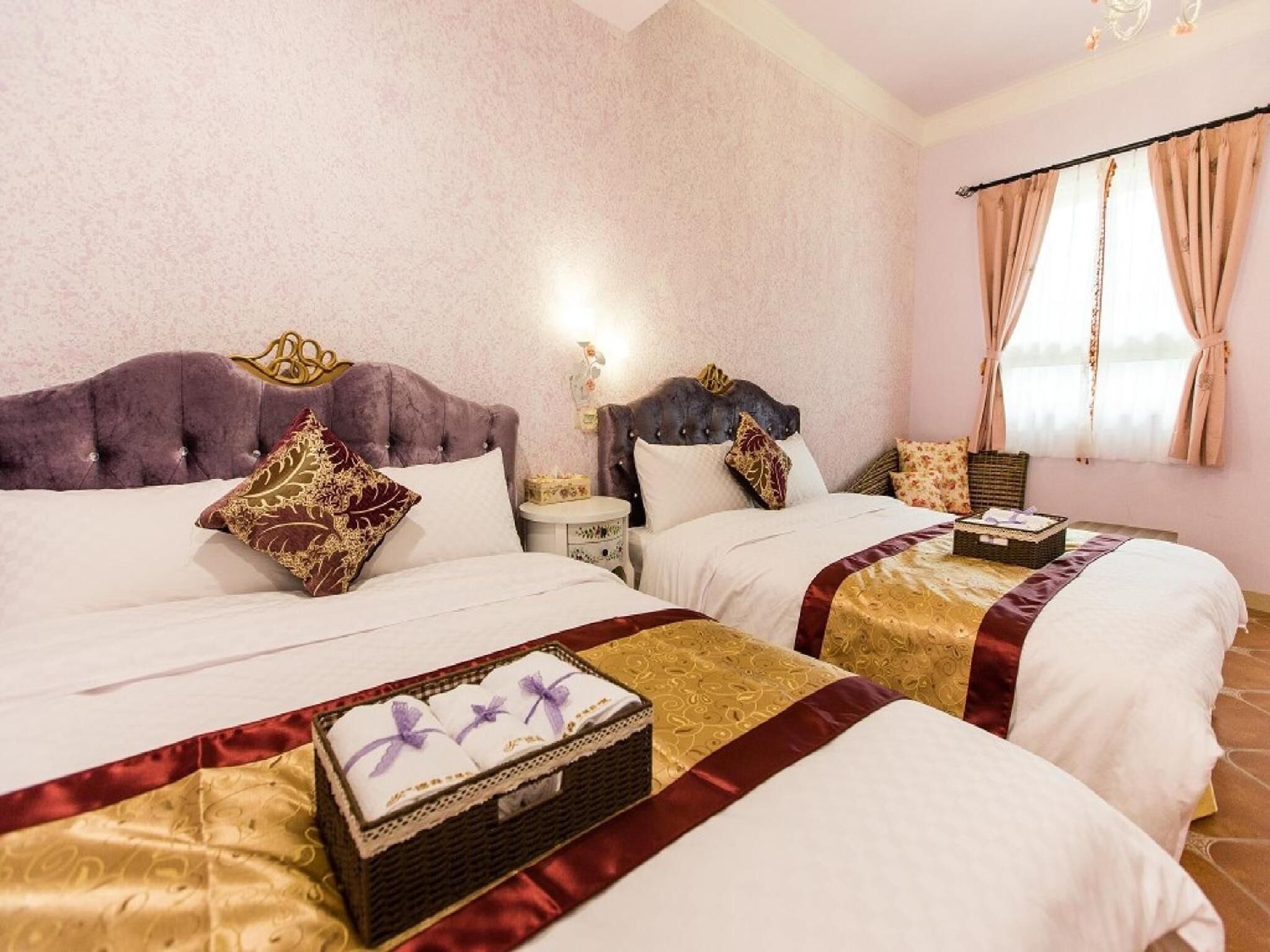 Quarto de hotel com duas camas, cada uma com cabeceira, mesa de cabeceira e mesa lateral. Há uma janela com cortinas, e um candelabro é visível no teto.