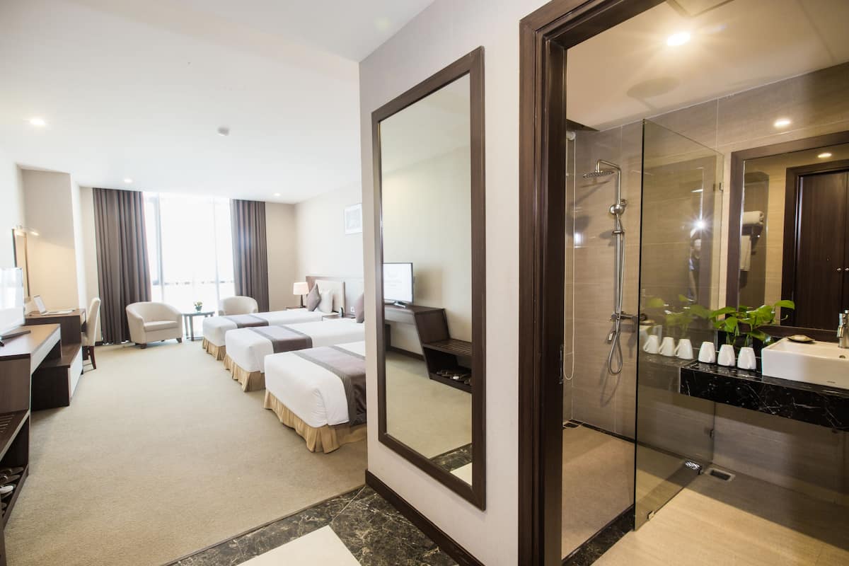 A hotel room with a large bed, a desk, a chair, a small table, a mirror, a shower area, and a bathroom with a sink and mirror.