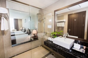 A modern hotel bathroom with a glass-enclosed shower, a large mirror, a marble sink, and a bedside table with a lamp. - Muong Thanh Grand Hoang Mai Hotel (Hoang Mai)