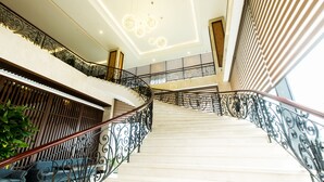 A grand staircase with ornate black railings and a wooden handrail, leading to a second-floor landing with a blue sofa and a potted plant.