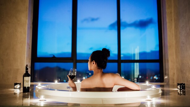 A person in a bathtub with a bottle of wine and candles, with a large window in the background.