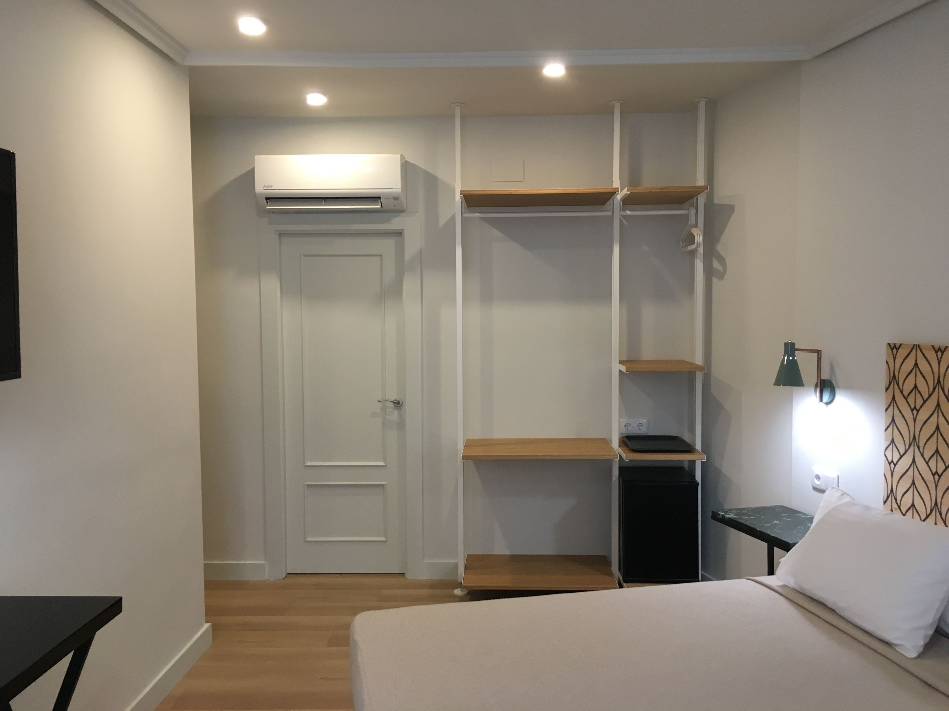 A bedroom with a bed, a wardrobe with shelves, a desk with a lamp, and an air conditioning unit.