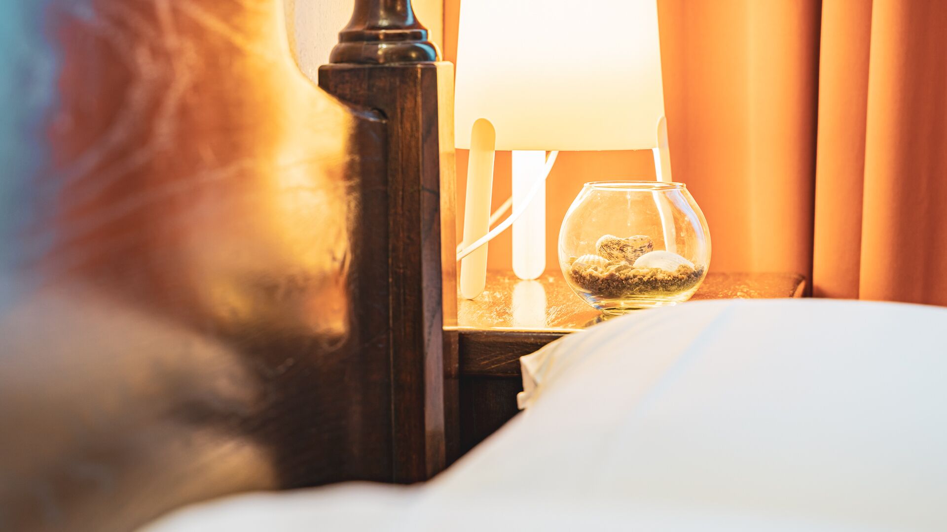 A bed with white bedding, a wooden nightstand with a lamp, and a glass containing seashells.