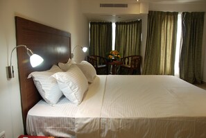 A hotel room with a large bed, two bedside lamps, a small table with chairs, and a window with curtains.