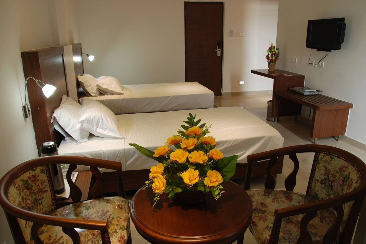 A hotel room with two beds, a desk, a chair, a TV, and a flower arrangement.