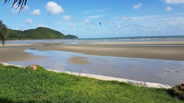 Una playa con una zona verde en primer plano, arena y una colina a lo lejos.