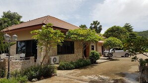 Una casa con techo de tejas, paredes de piedra y un pequeño jardín con diversas plantas y un coche blanco estacionado en la entrada.