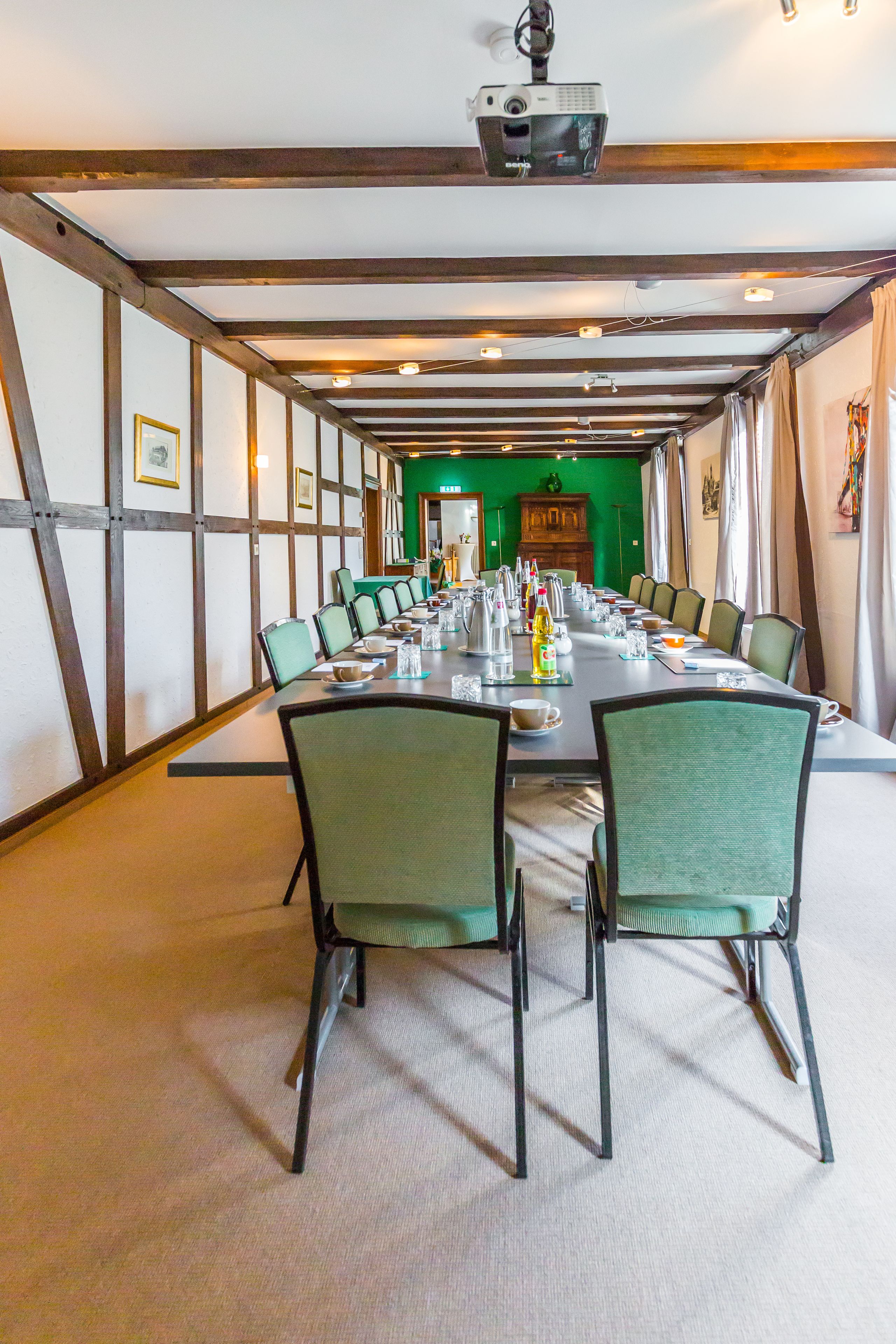 Un salón de conferencias con una larga mesa dispuesta para una reunión, rodeada de sillas y un proyector montado en el techo.