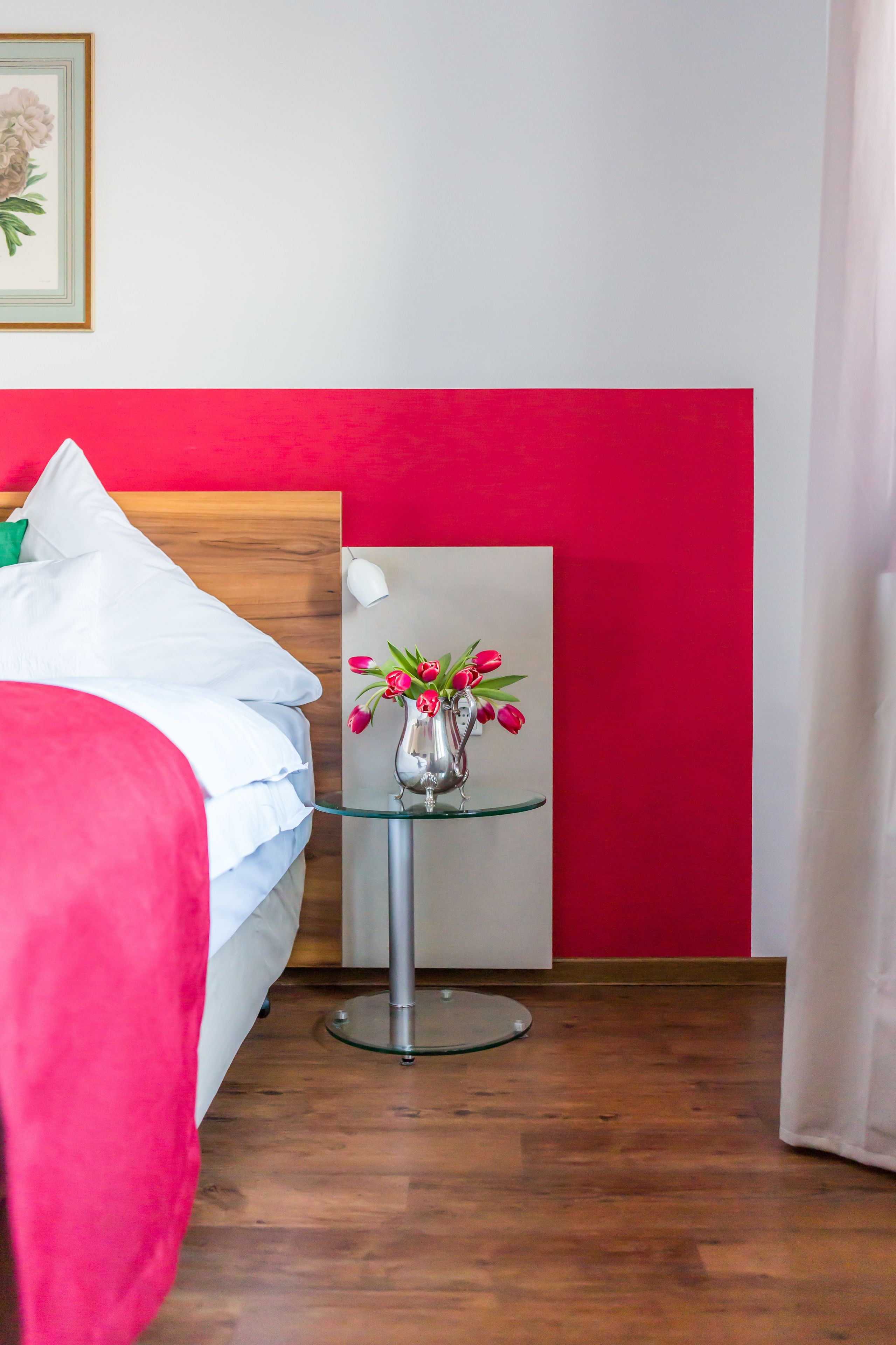 Quarto de hotel com cama, mesa de vidro com um vaso de flores, parede com detalhes em vermelho e um quadro na parede.