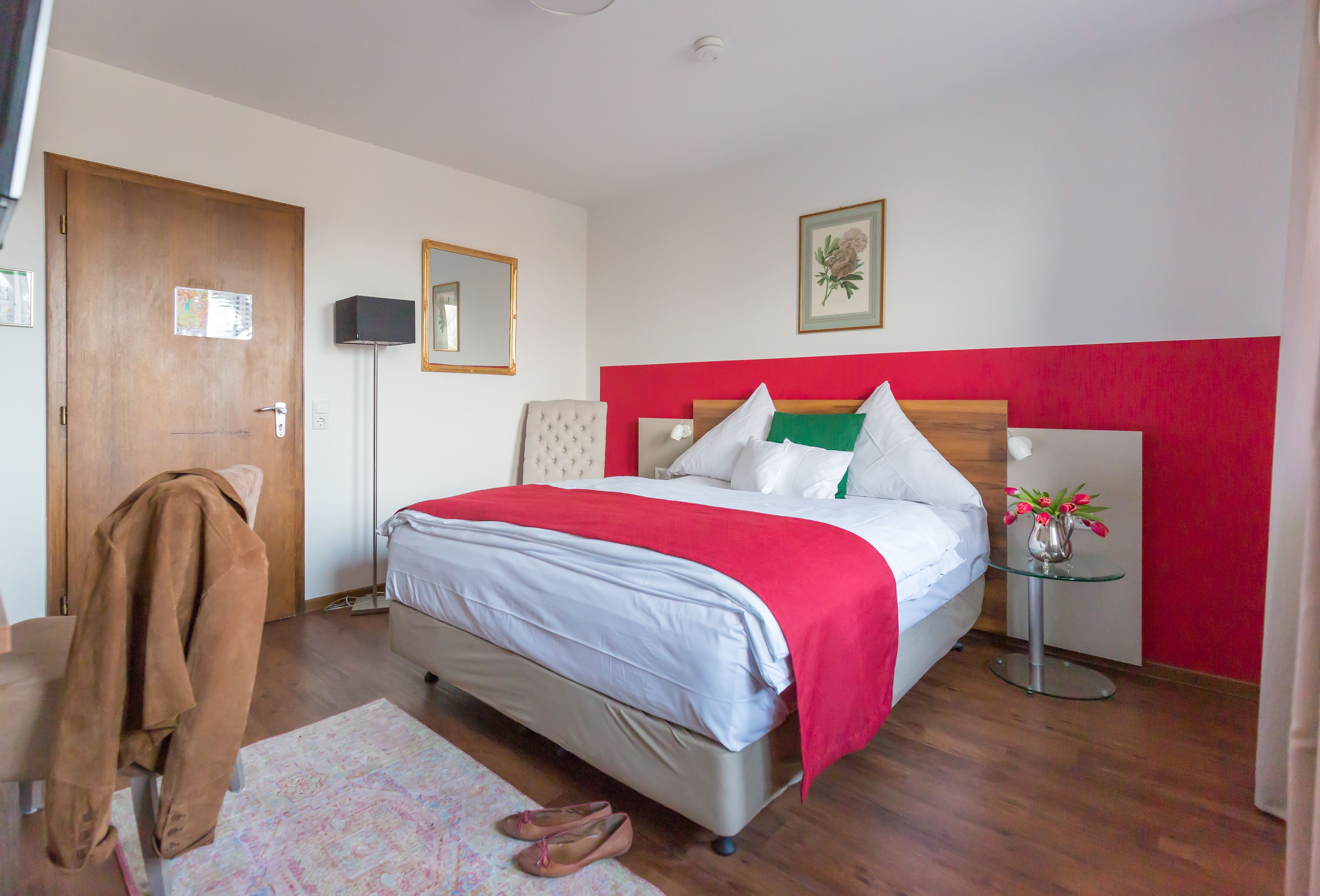 A hotel room with a bed, a chair, a small table with a vase of flowers, a mirror, and a door.
