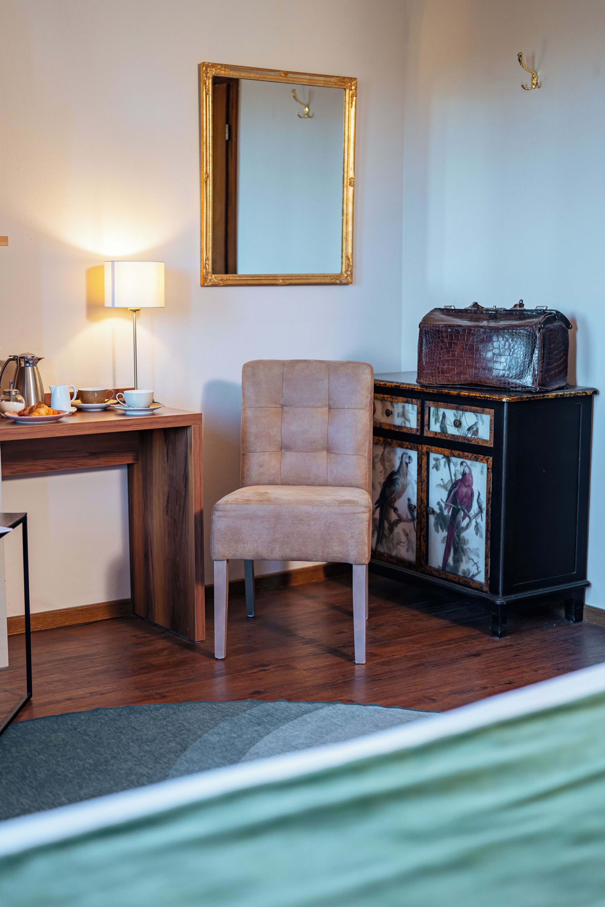 Una habitación con un escritorio de madera, una silla, una lámpara, un armario con obras de arte y una cama.