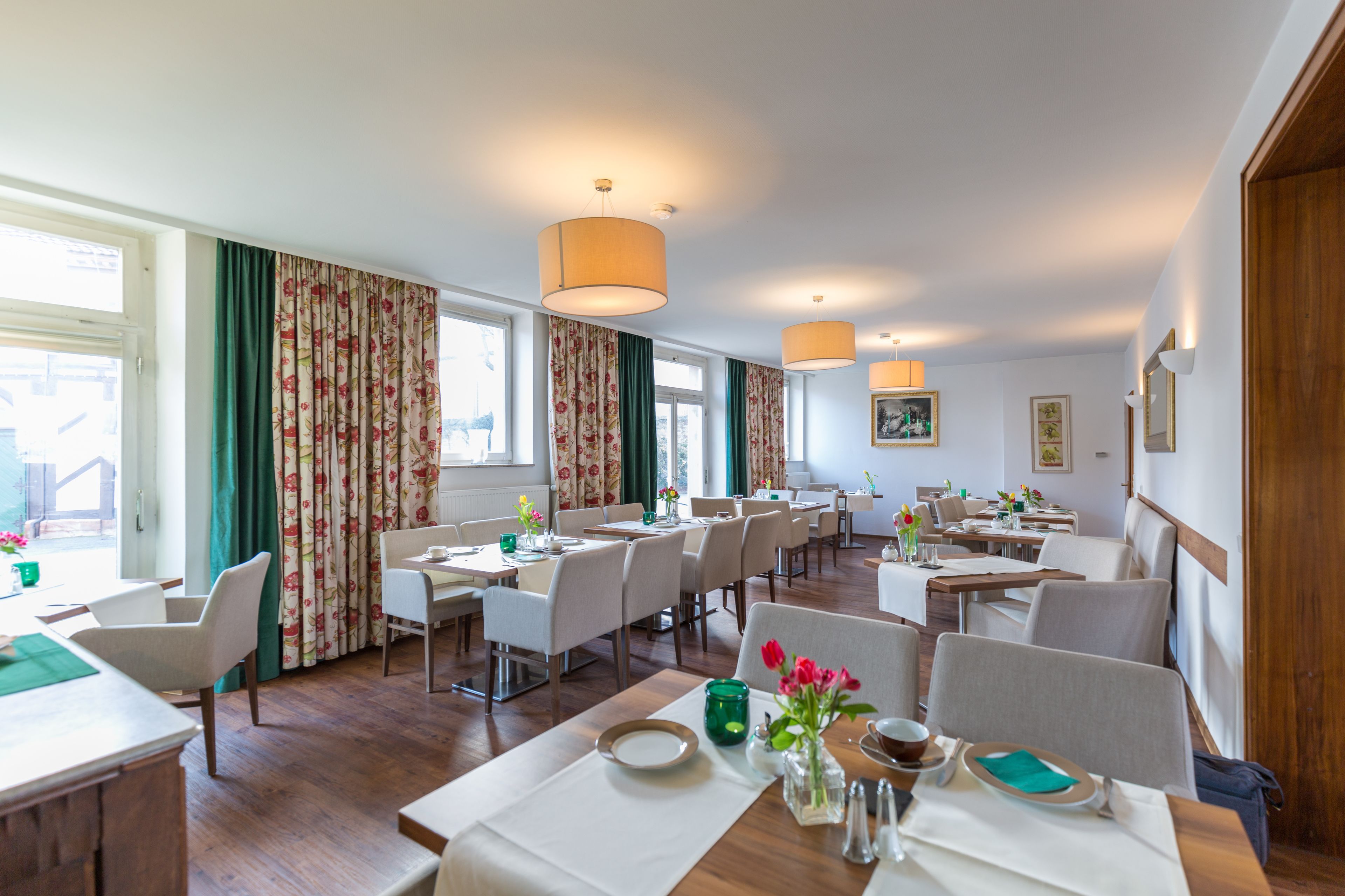 A spacious hotel dining area with multiple tables and chairs, large windows with curtains, and hanging lamps.