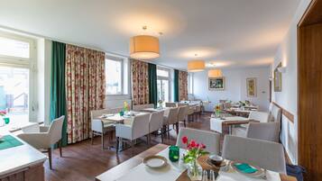 A spacious hotel dining area with multiple tables and chairs, large windows with curtains, and hanging lamps.