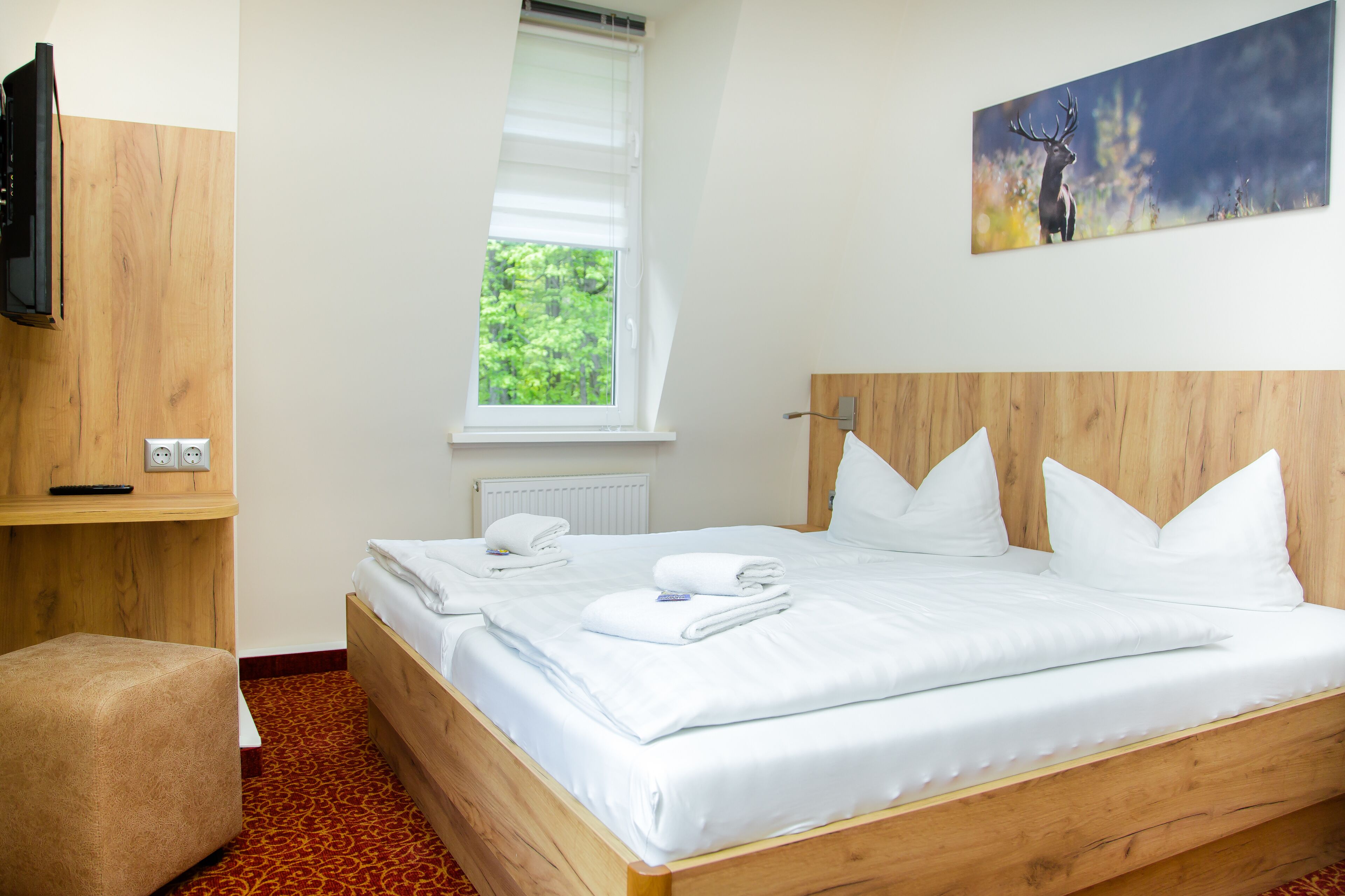 Een netjes opgemaakt bed met witte lakens, een houten hoofdbord, een klein nachtkastje met een afstandsbediening, een raam met zonwering, en een schilderij van een hert aan de muur.