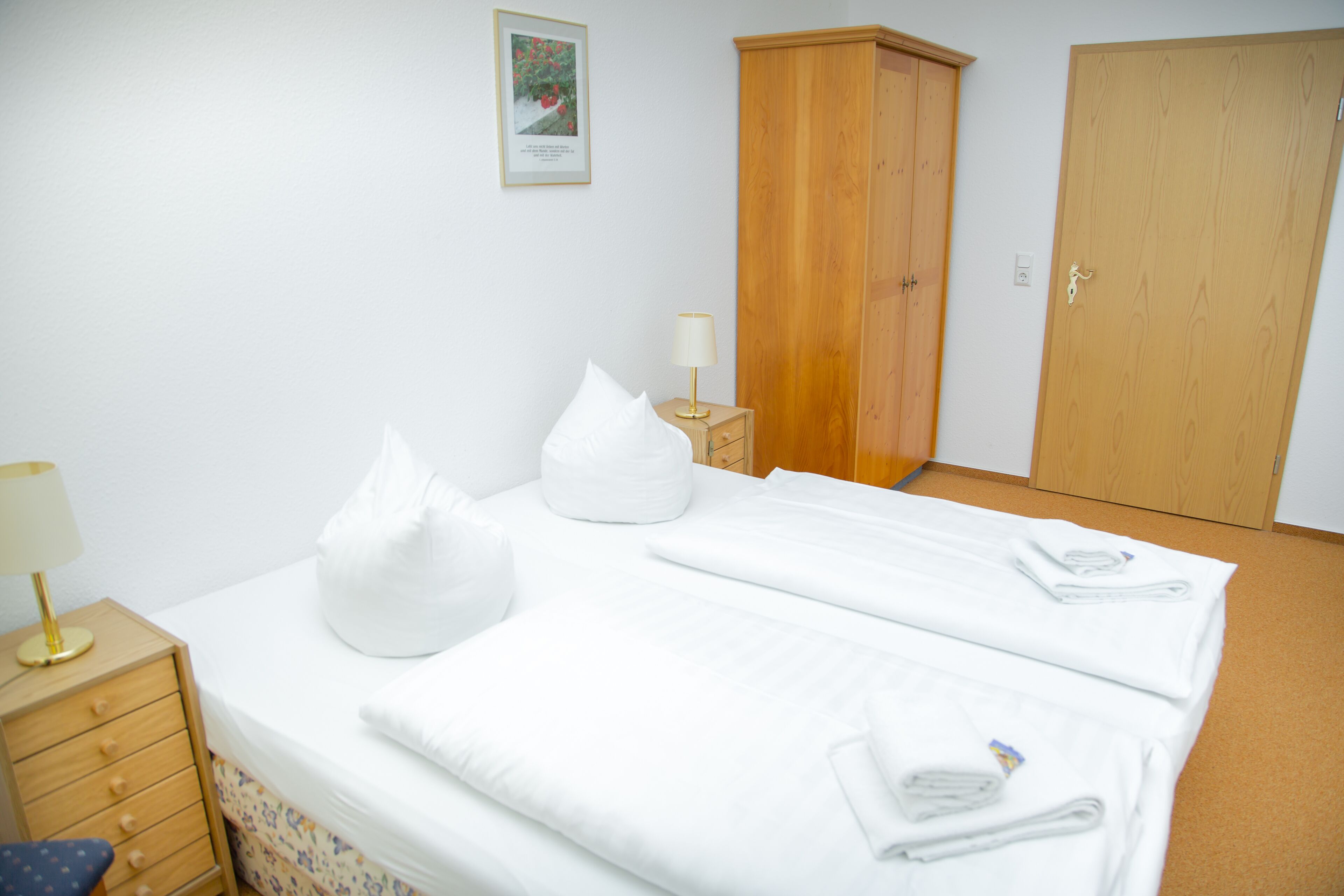 A neatly made bed with pillows and folded towels, a wooden wardrobe, a bedside table with a lamp, and a framed picture on the wall.