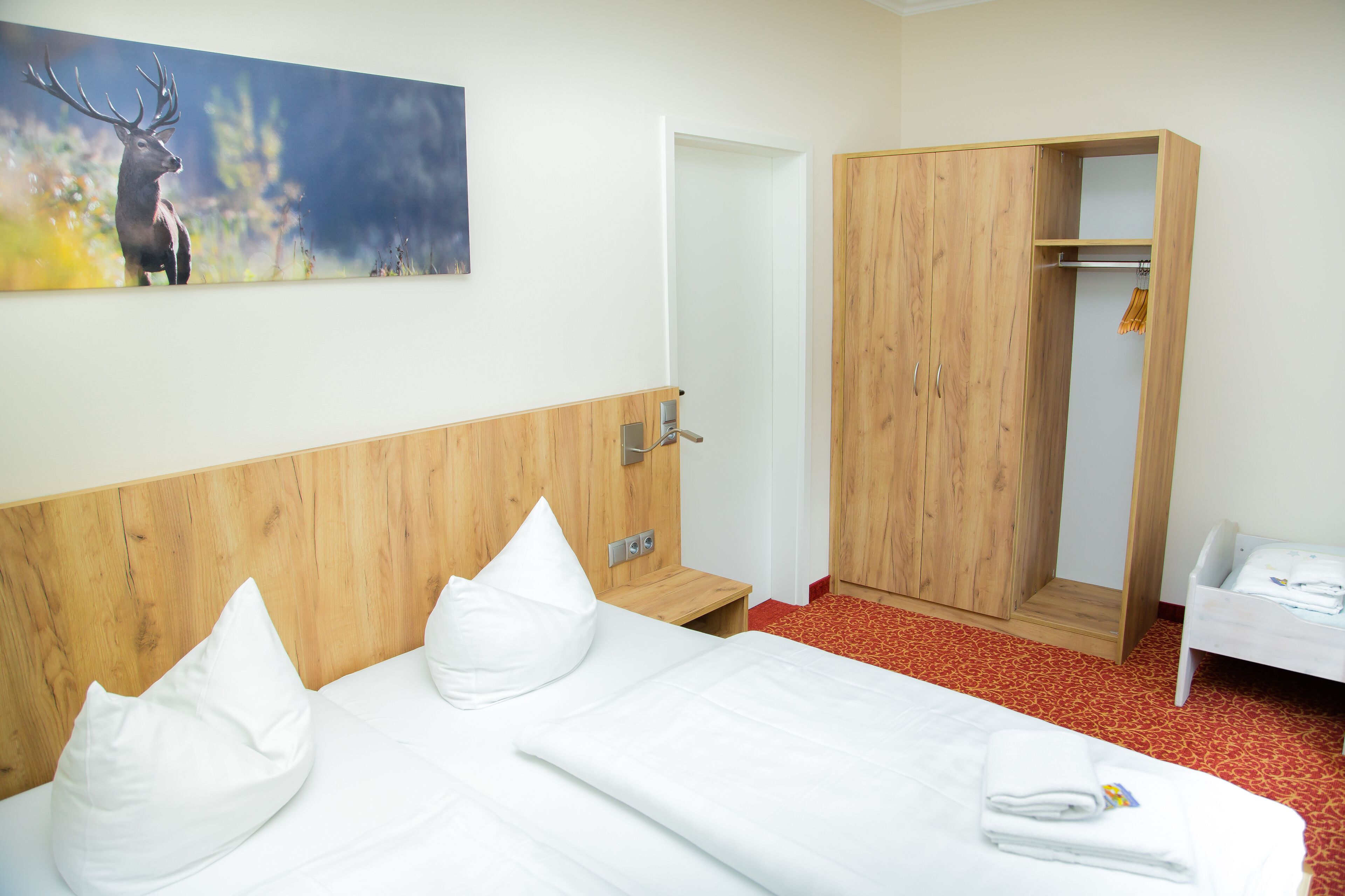 A hotel room with a wooden headboard, a bed with white linens, a wooden wardrobe, and a painting of a deer on the wall.